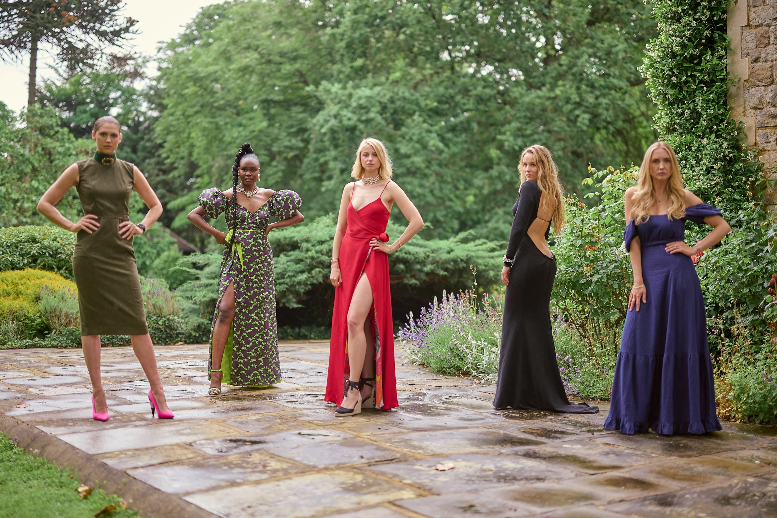 Five women wearing formal dresses standing outdoors on a stone patio with green trees and shrubs in the background.