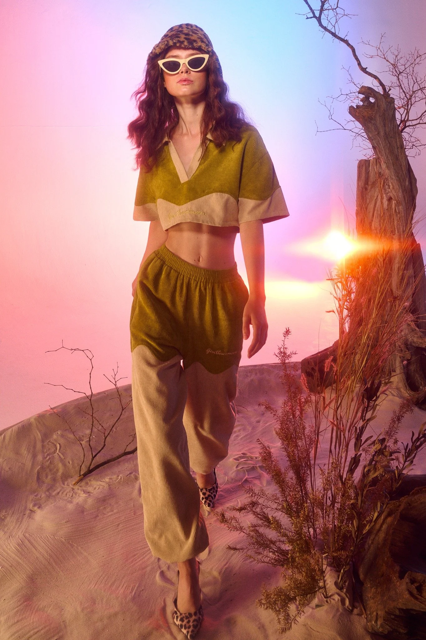 A woman dressed in retro-style clothing walking on a sandy landscape with a leafless tree and plants at sunset, wearing sunglasses and a hat.