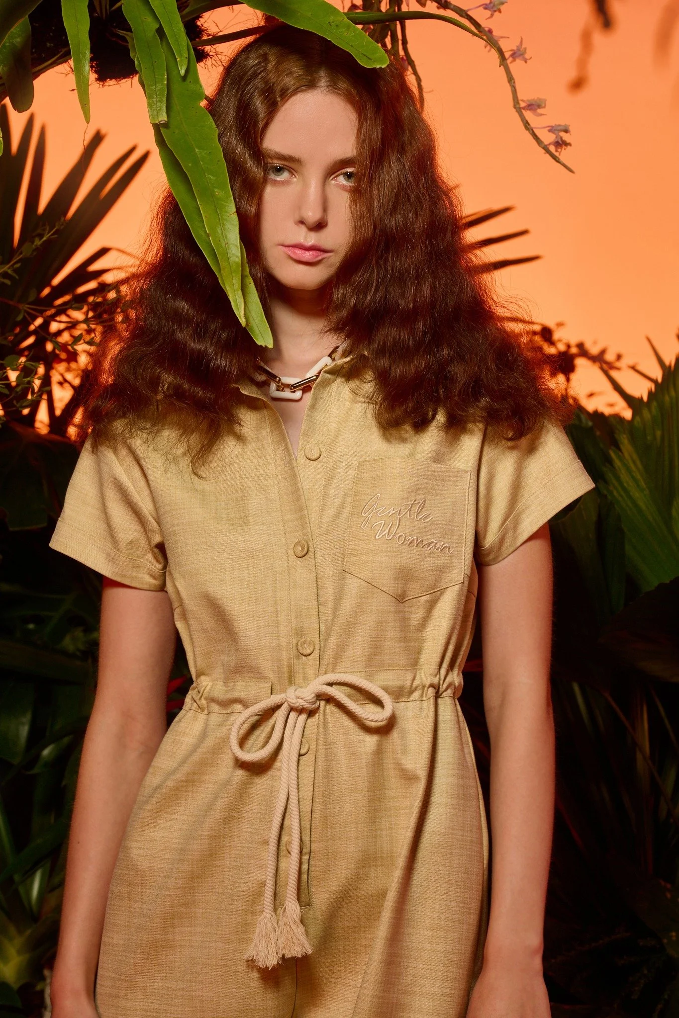 Young woman with long, curly brown hair wearing a beige jumpsuit with a tied waist and embroidered pocket, standing among tropical plants with a sunset or warm-colored background.