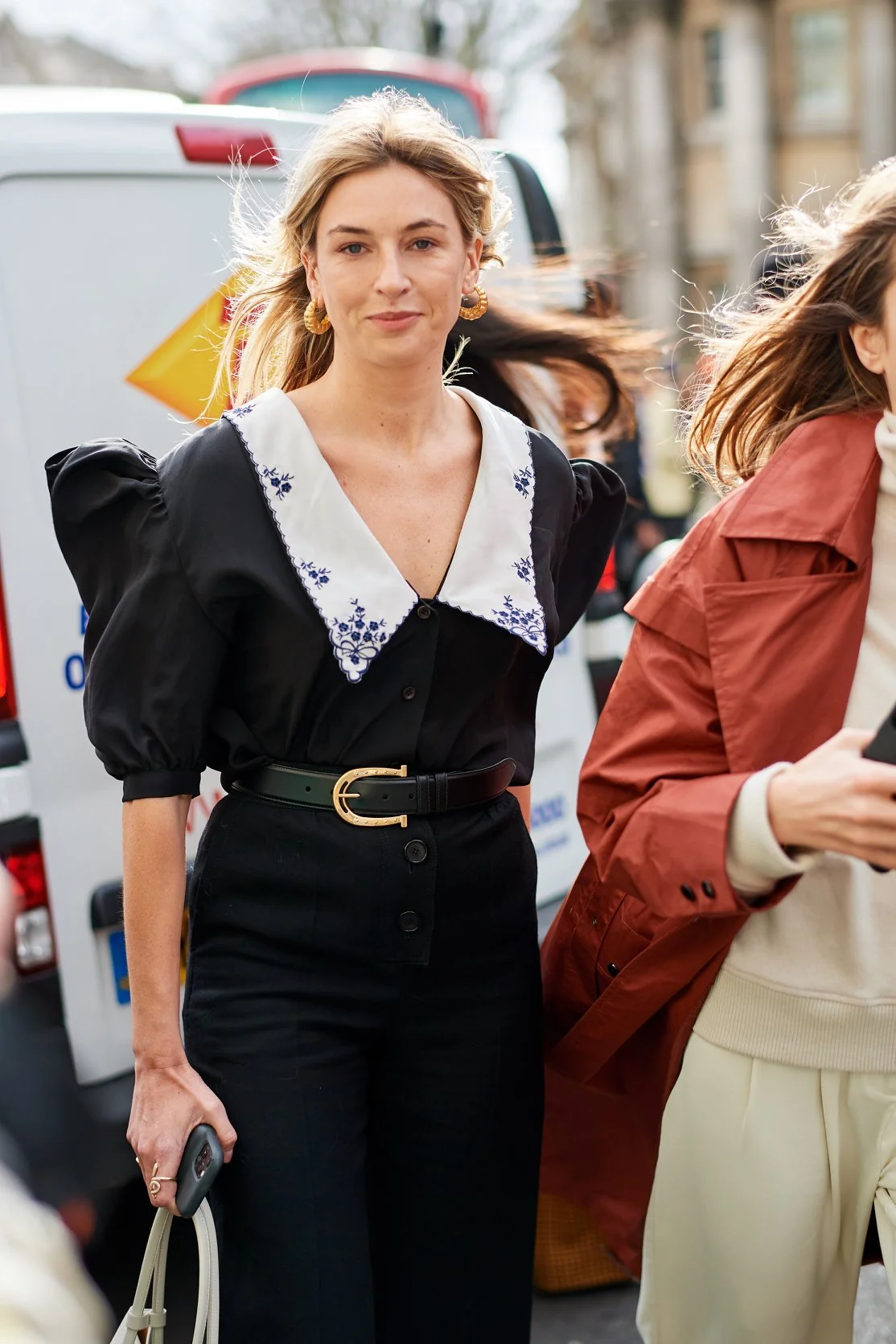 A woman with blonde hair wearing a black blouse with puffed sleeves and a large white collar with blue embroidery, high-waisted black pants with a belt, holding a phone and a beige purse in an outdoor urban setting.