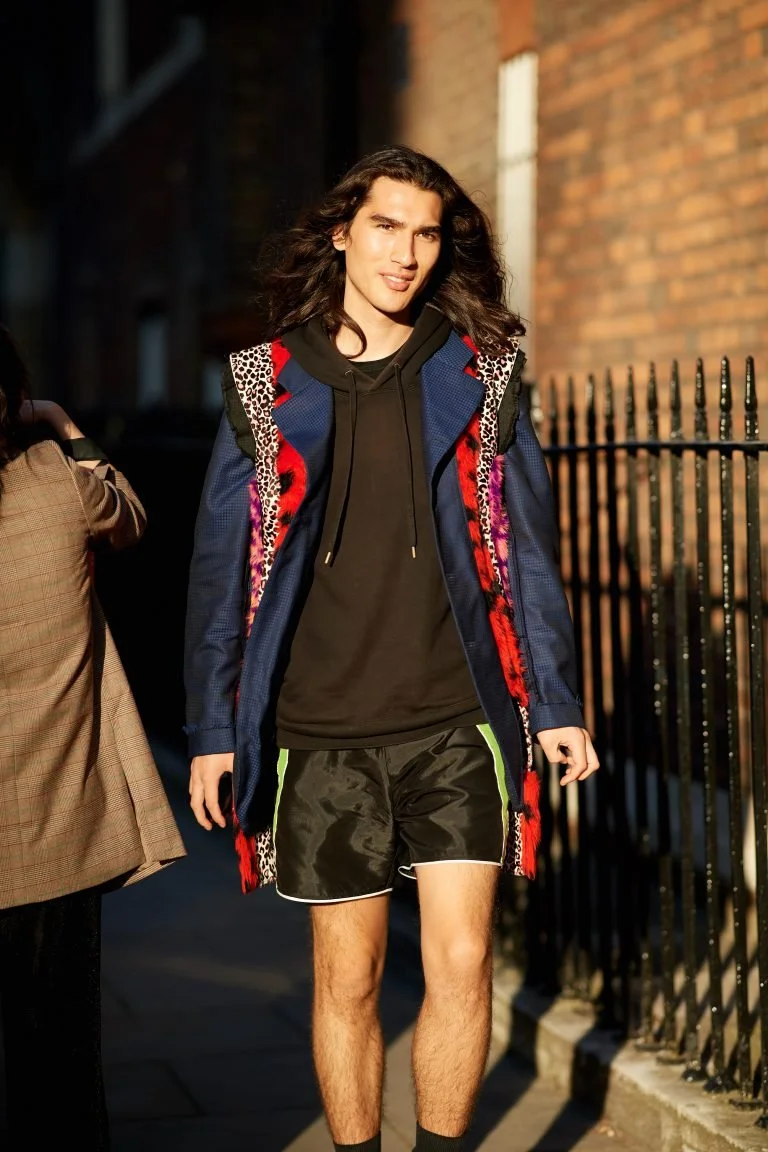Young man with long hair wearing a colorful jacket, black hoodie, and black shorts with green trim, walking outdoors on a sunny day.