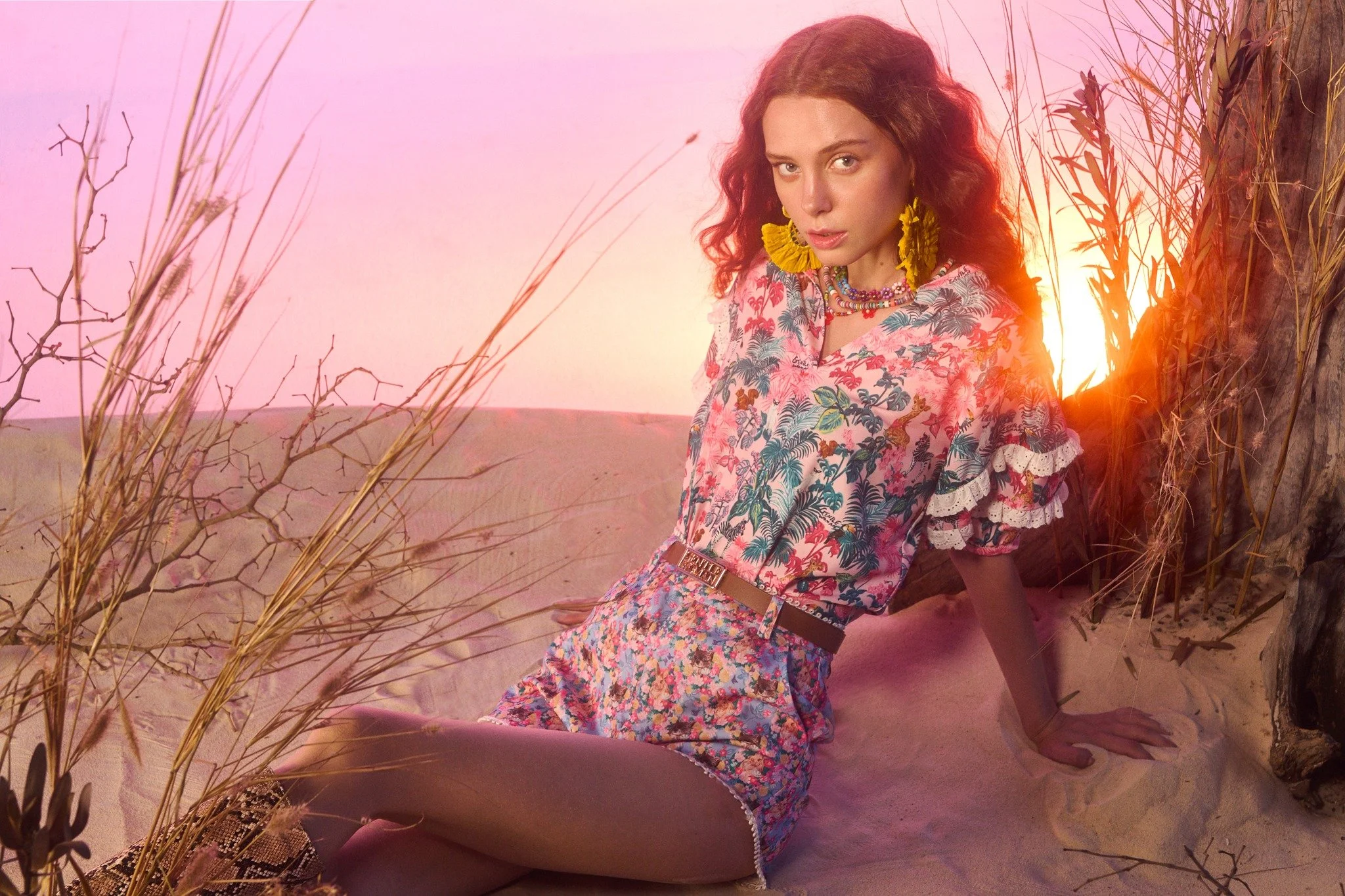A woman with curly red hair sitting on the sand in a desert, surrounded by dry plants and a tree, with a sunset in the background illuminating the scene with warm light. She is wearing colorful floral clothing, large yellow earrings, and layered neck