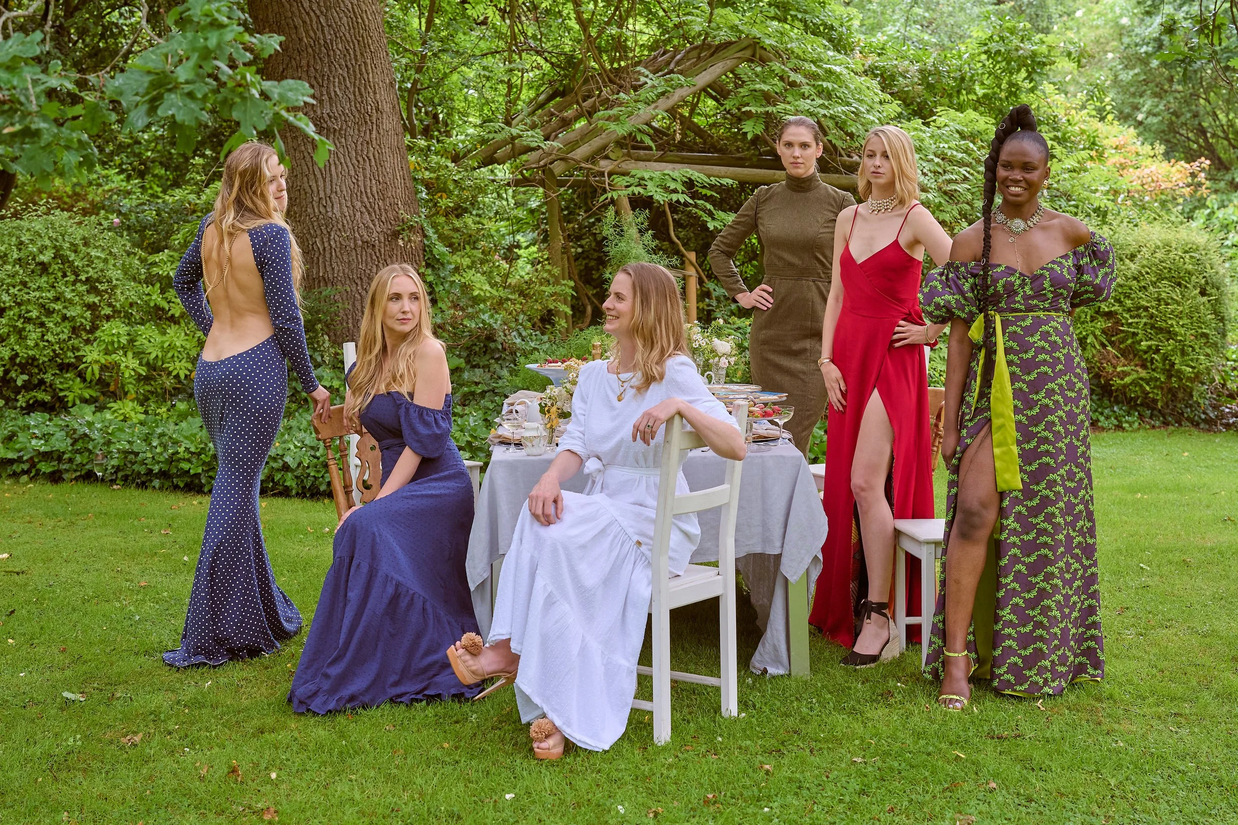 Seven women are gathered outdoors around a garden table dressed in colorful elegant dresses. One woman in a white dress sits on a chair, three women stand to the right, one woman is seated, and three women stand on the left side of the table. The set