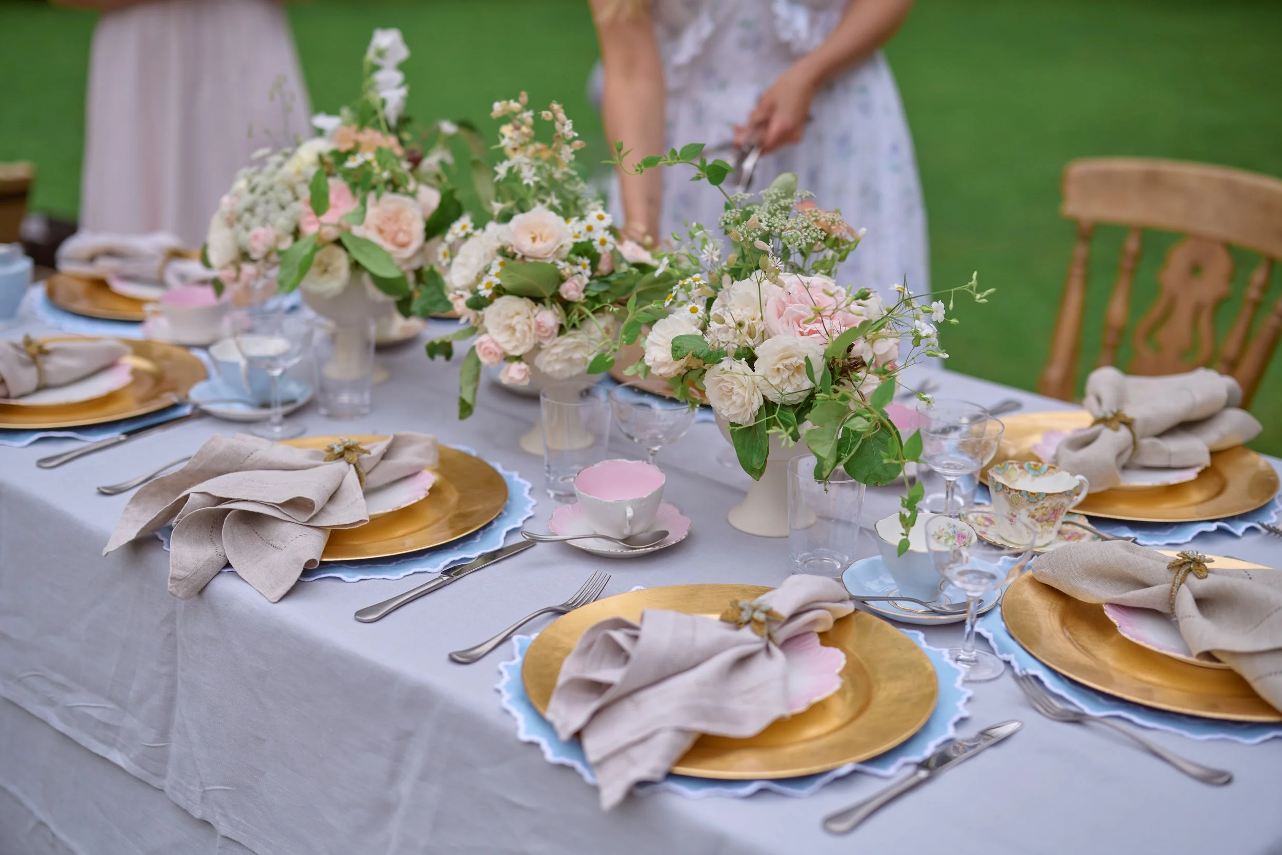Elegant outdoor dining table decorated with white and pink floral arrangements, gold chargers, beige napkins tied with gold rings, and assorted glassware on a white tablecloth.
