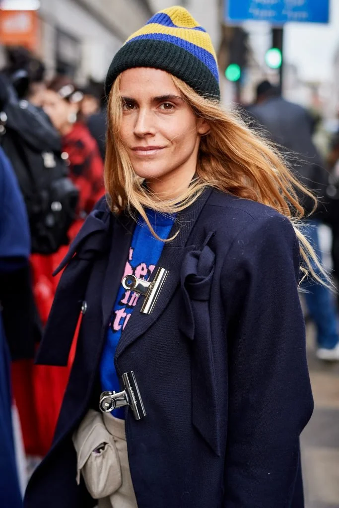 A woman with long blonde hair wearing a blue and yellow beanie, a dark coat with decorative clips, and a blue shirt, standing outdoors in a crowd.