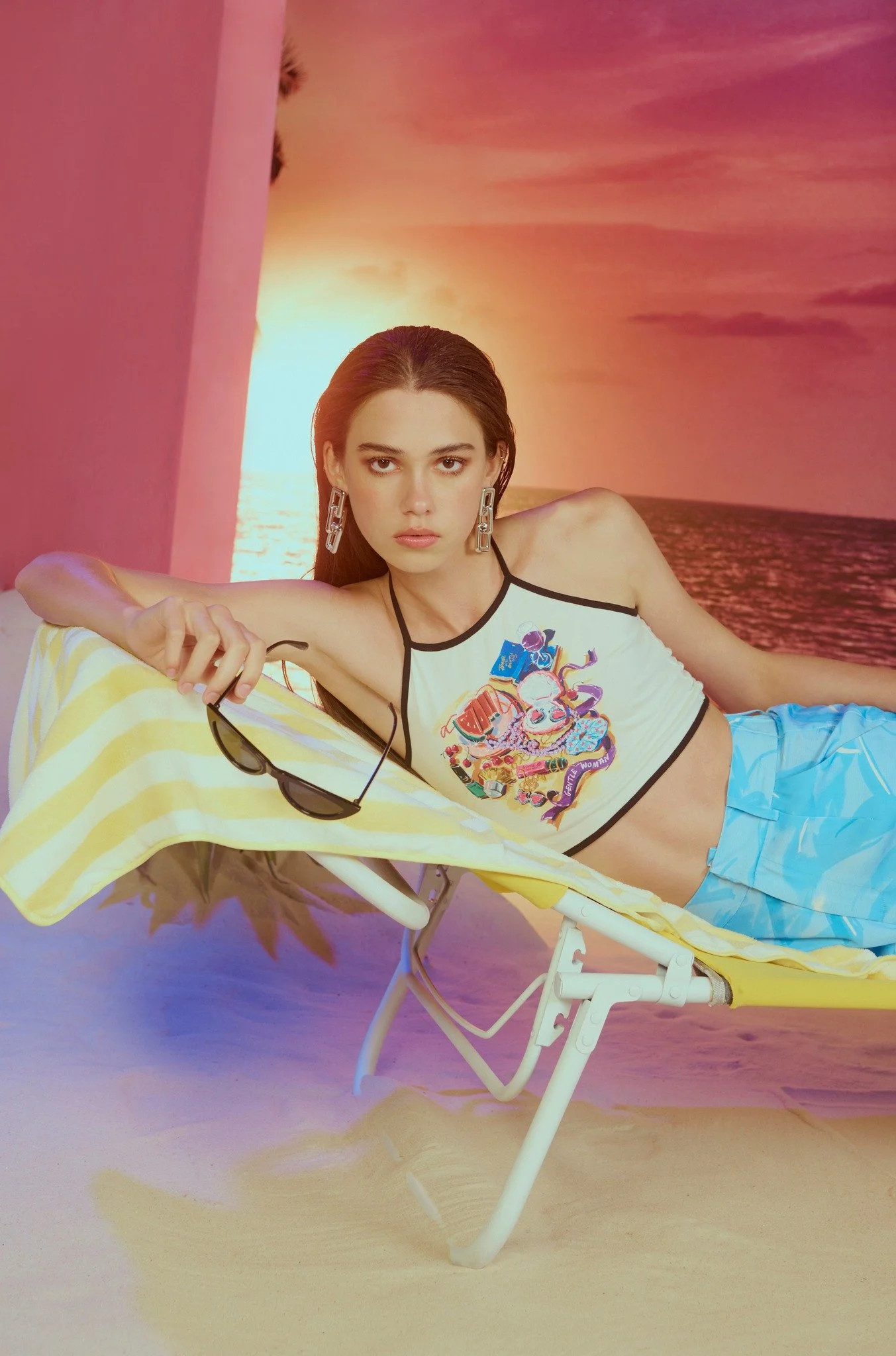 A young woman with long dark hair lies on a yellow lounge chair on a sandy beach with a sunset in the background. She wears large earrings, a sleeveless crop top with colorful graphic print, and light blue shorts. She holds sunglasses in her right ha