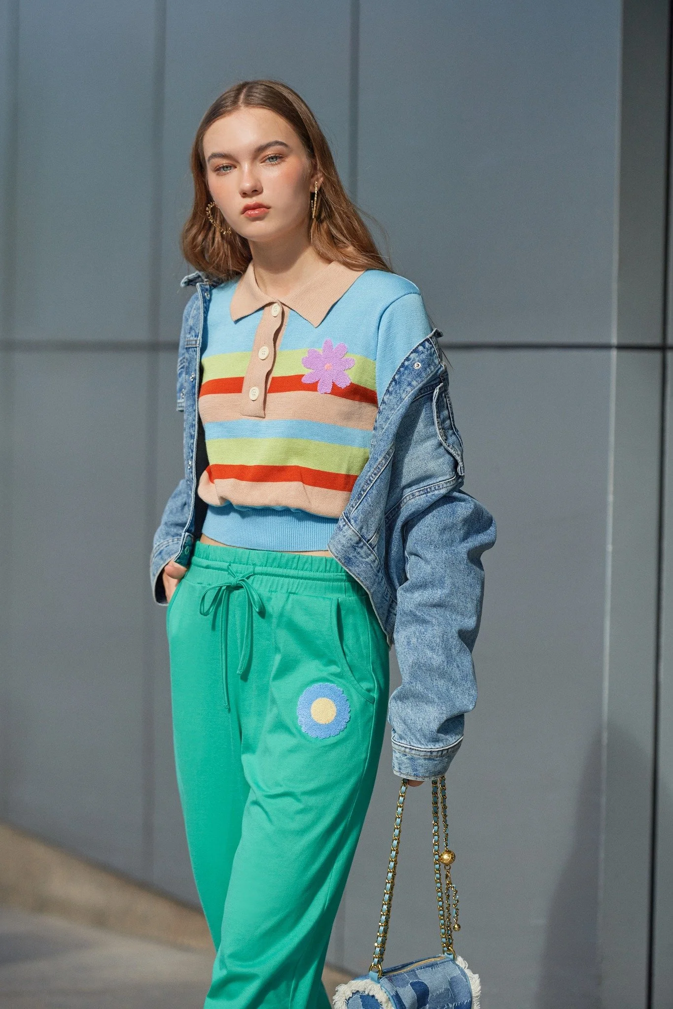 A young woman with long hair, wearing a colorful striped shirt with a purple flower detail, a denim jacket, and green pants with a daisy on the pocket, holding a chain-strap purse, standing against a gray wall.