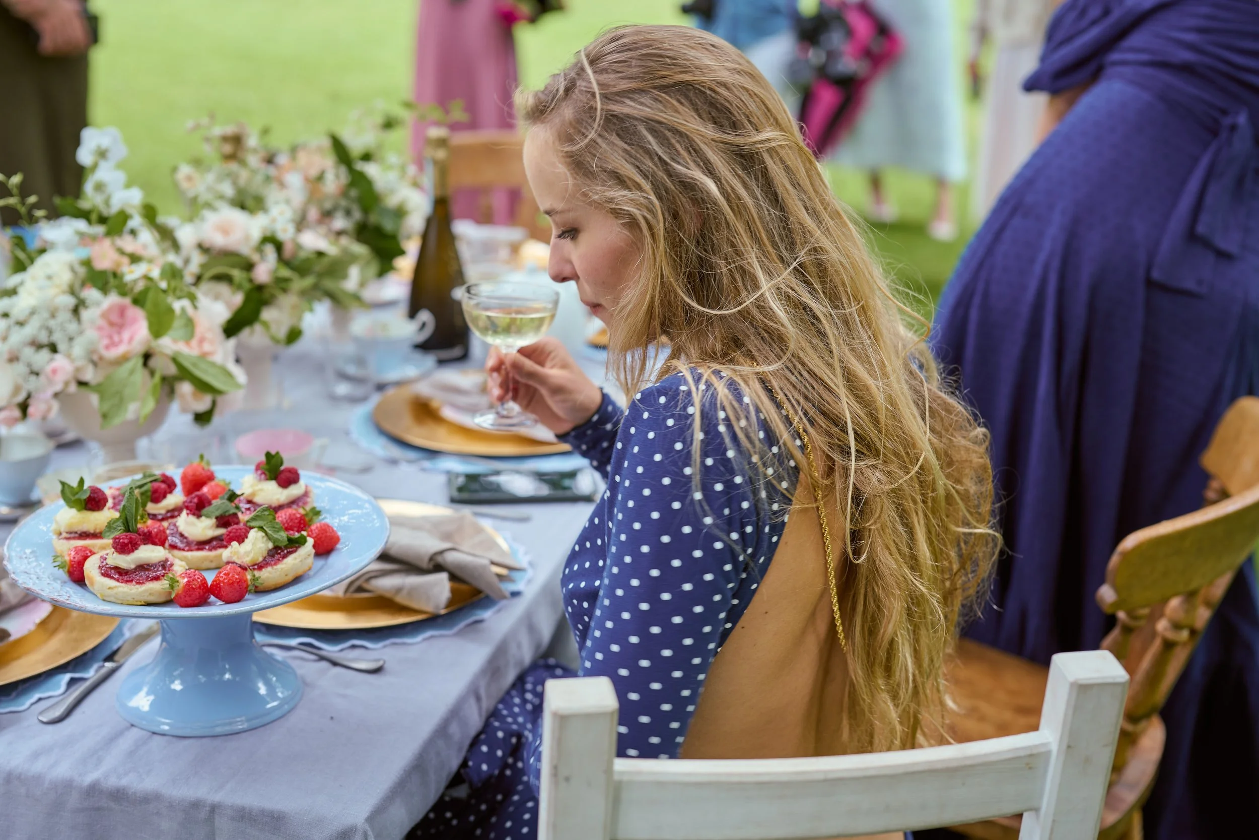 A young woman with long, curly blonde hair is sitting at a table with a light gray tablecloth, holding a glass of white wine or champagne, with dessert on a blue cake stand nearby. The table is decorated with flowers and set for a gathering or celebr