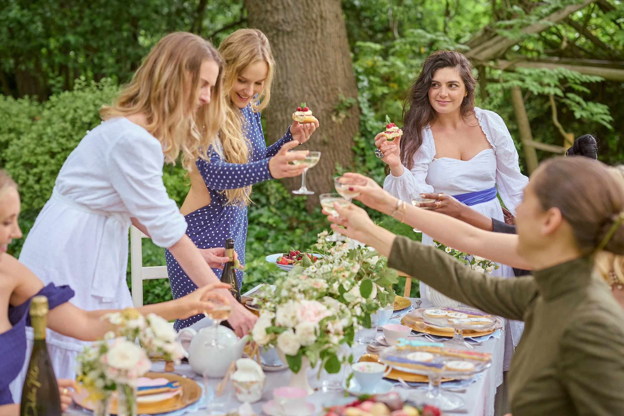 Group of women celebrating outdoors with cake and champagne on a decorated table surrounded by trees.