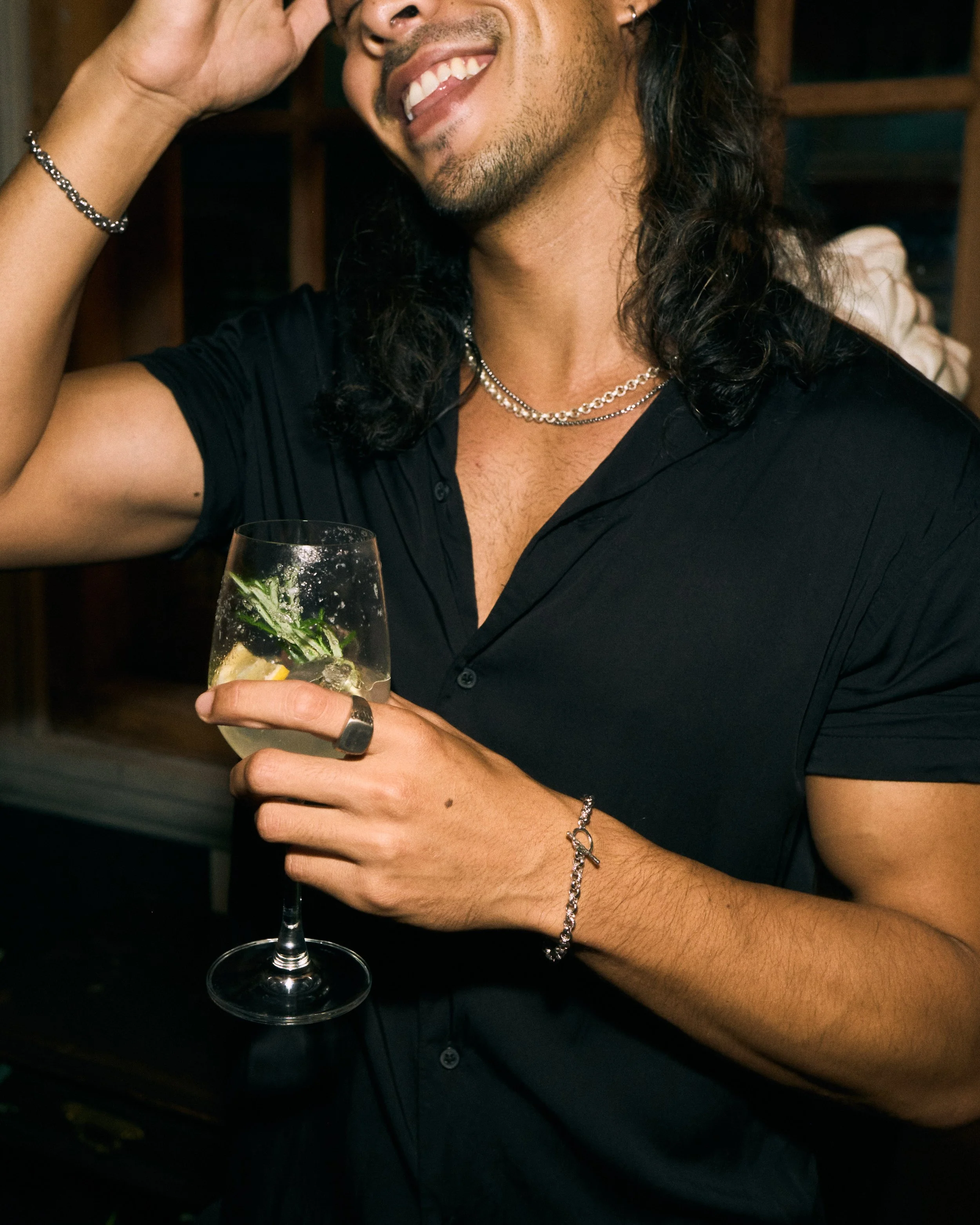 A man with long dark curly hair, smiling and holding a glass of what appears to be a cocktail with herbs and lemon slices. He is wearing a black shirt, silver jewelry, and accessories, and is seen in a cozy indoor setting.