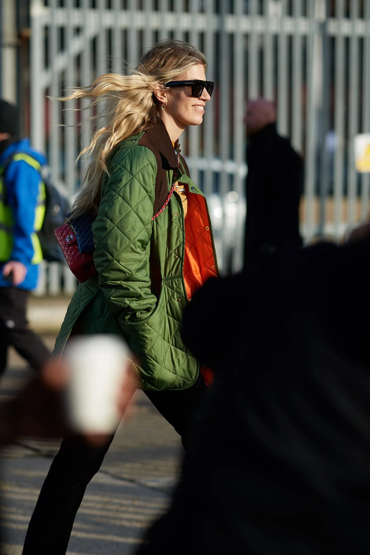A smiling woman with long blonde hair wearing a green quilted jacket, black sunglasses, and earrings walking outdoors.