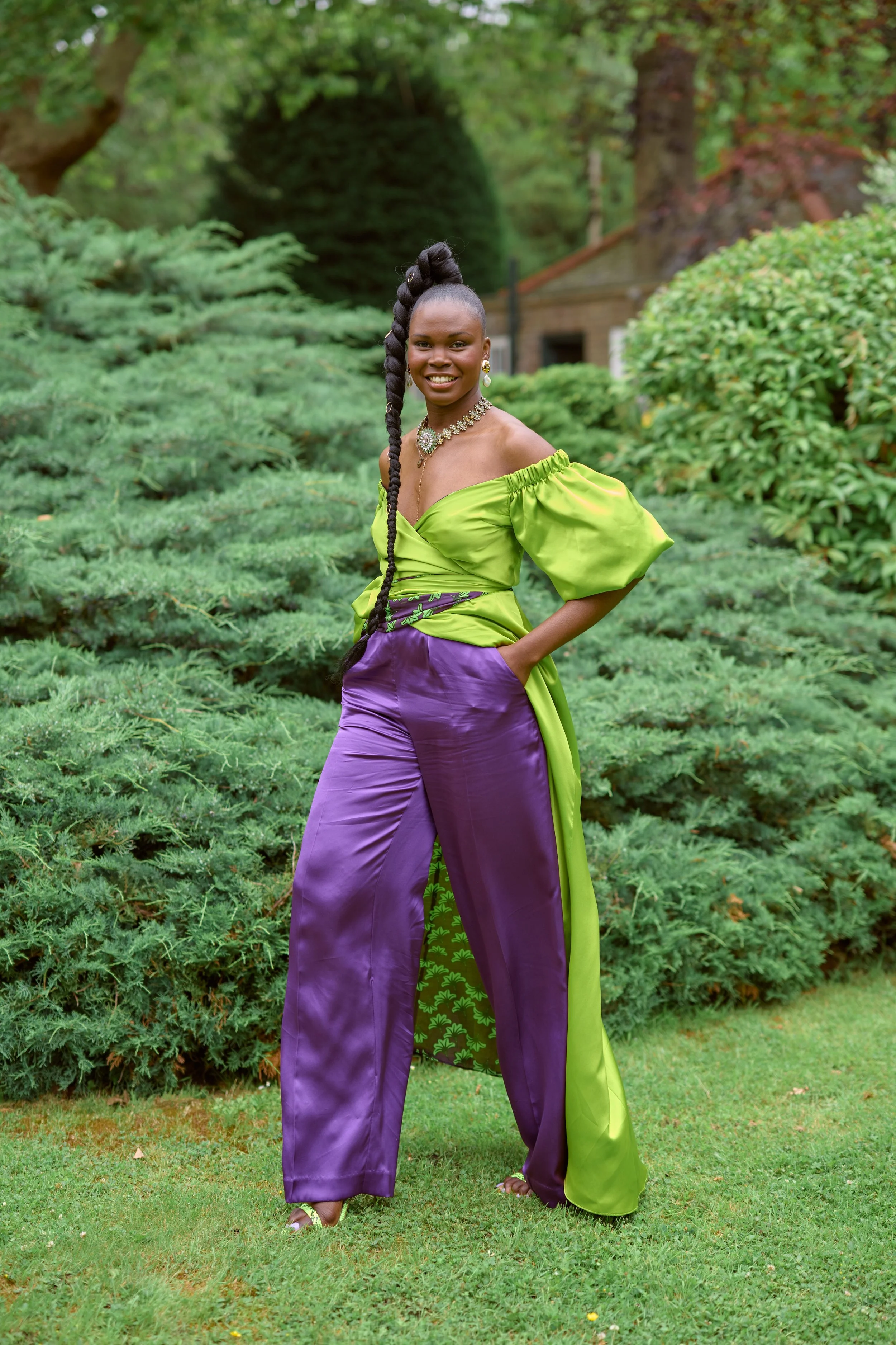 A woman wearing a vibrant green and purple dress with puffy sleeves, standing outdoors in a lush garden with green shrubs and trees, smiling.