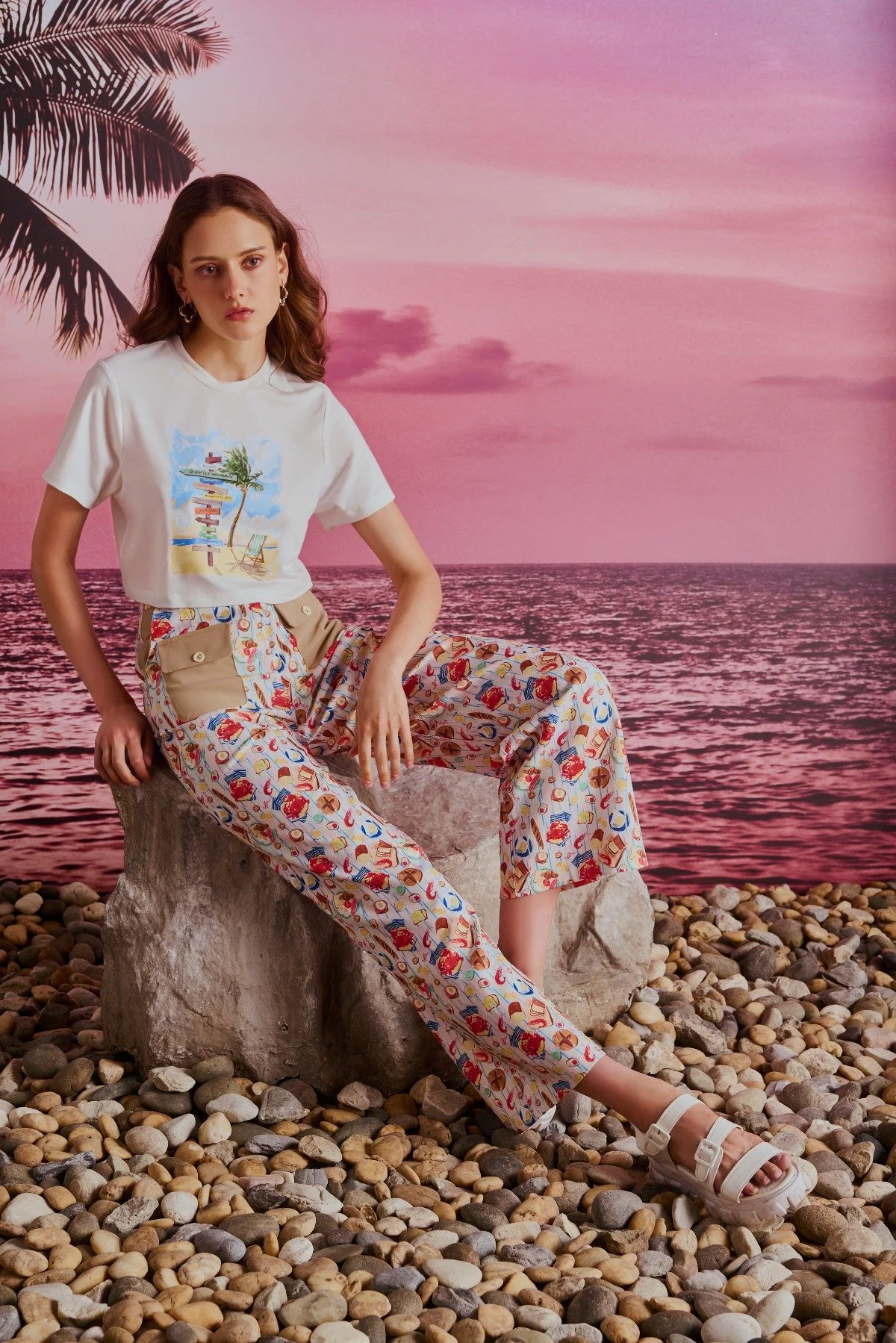 A young woman sitting on a large stone on a pebbled beach during sunset, with a tropical backdrop of pink sky, ocean, and palm trees, wearing a white graphic t-shirt and colorful patterned pants.