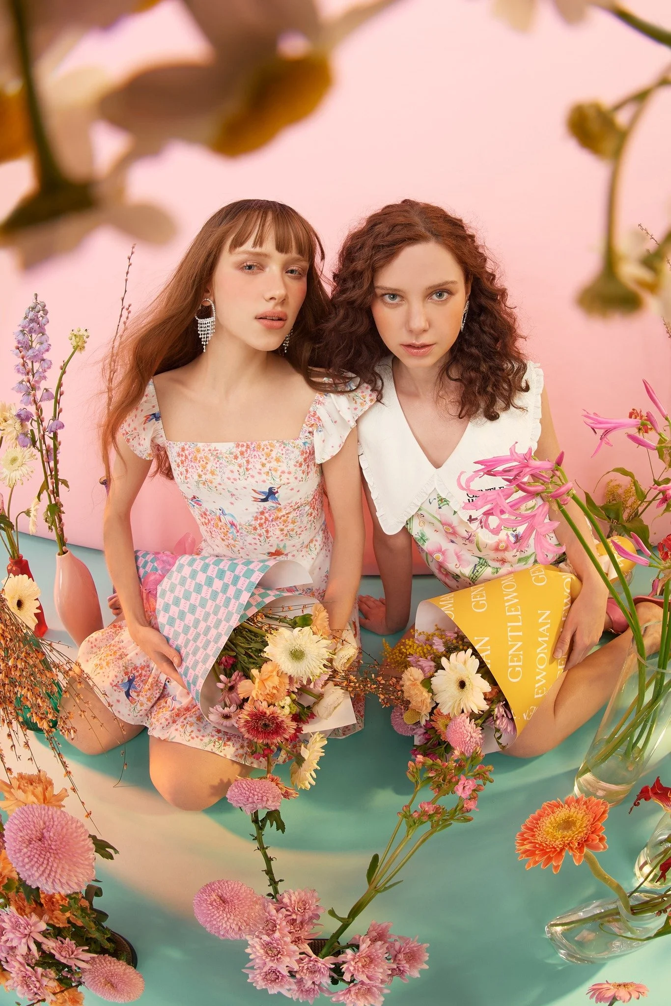 Two women sitting on the floor surrounded by various colorful flowers in vases, with pink and pastel-colored background.