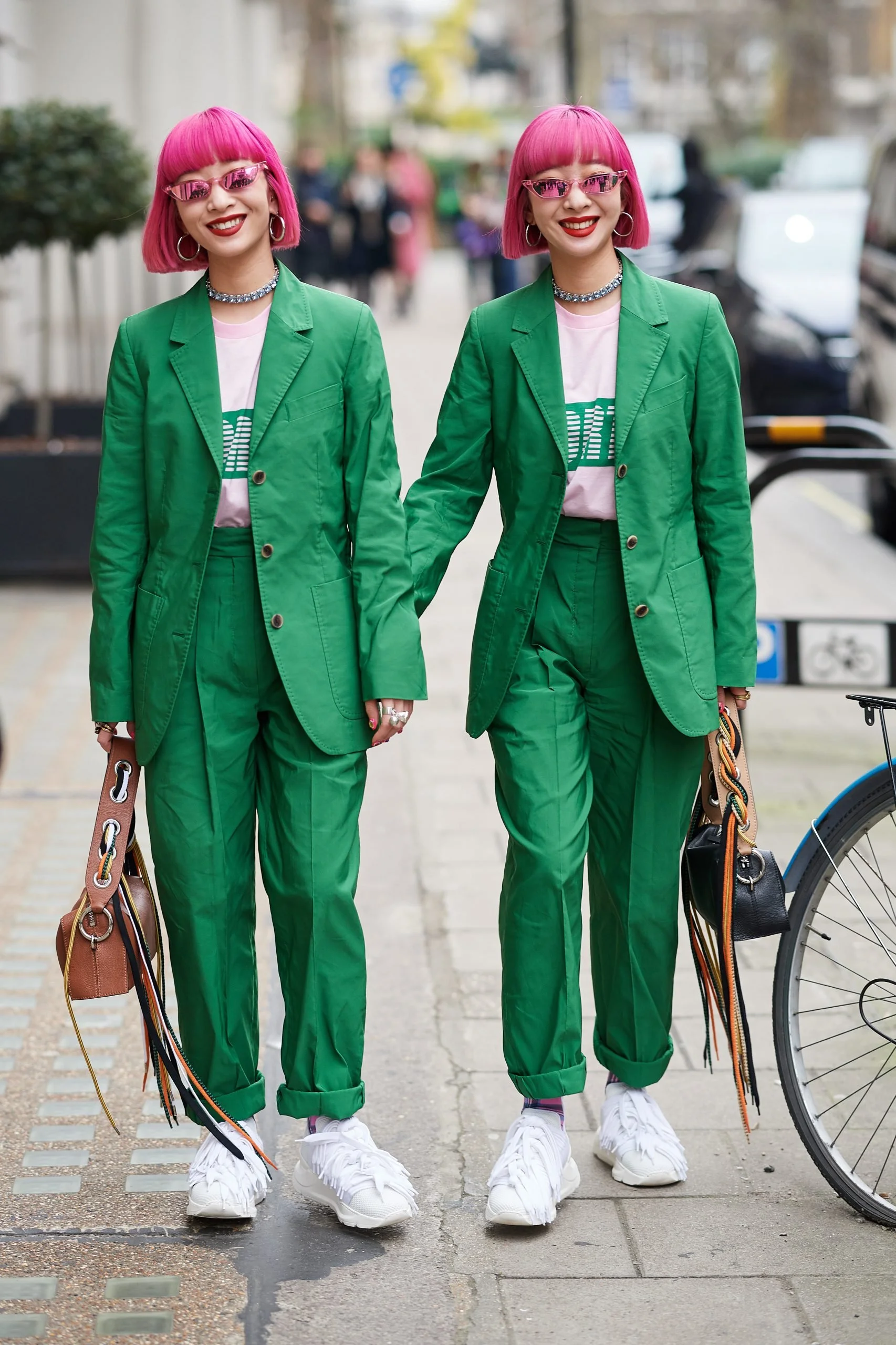 Two women with pink hair, wearing matching bright green suits, pink sunglasses, and white sneakers, walking together on a city street while holding hands and carrying handbags.