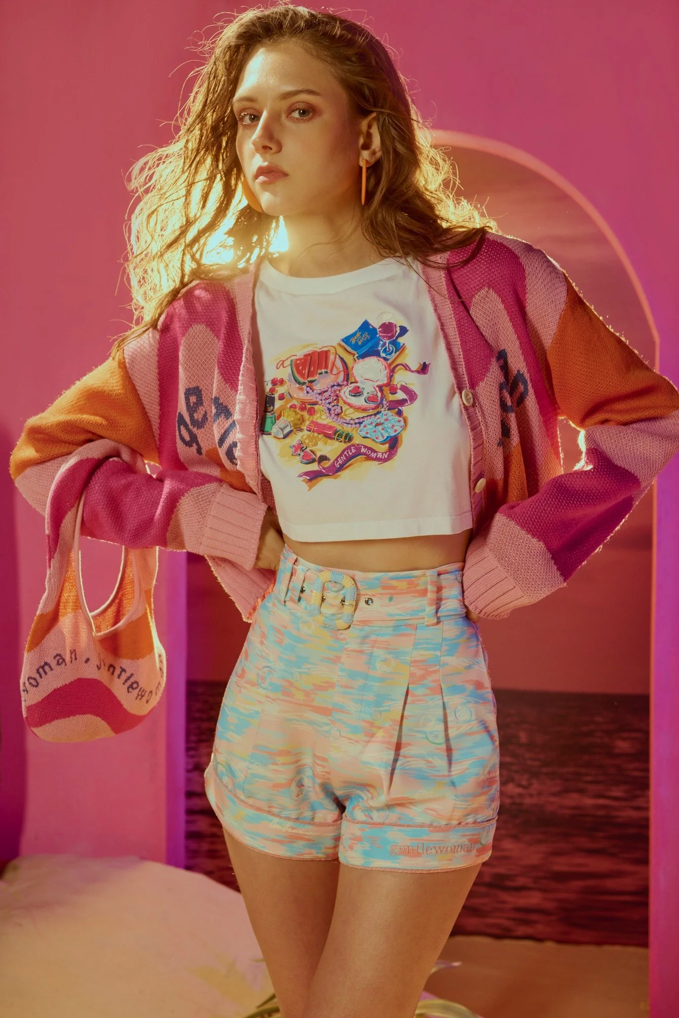 Young woman standing indoors with pink walls and a sunset beach scene in the background, wearing a colorful cardigan, a graphic crop top, pastel shorts, and carrying a matching tote bag.