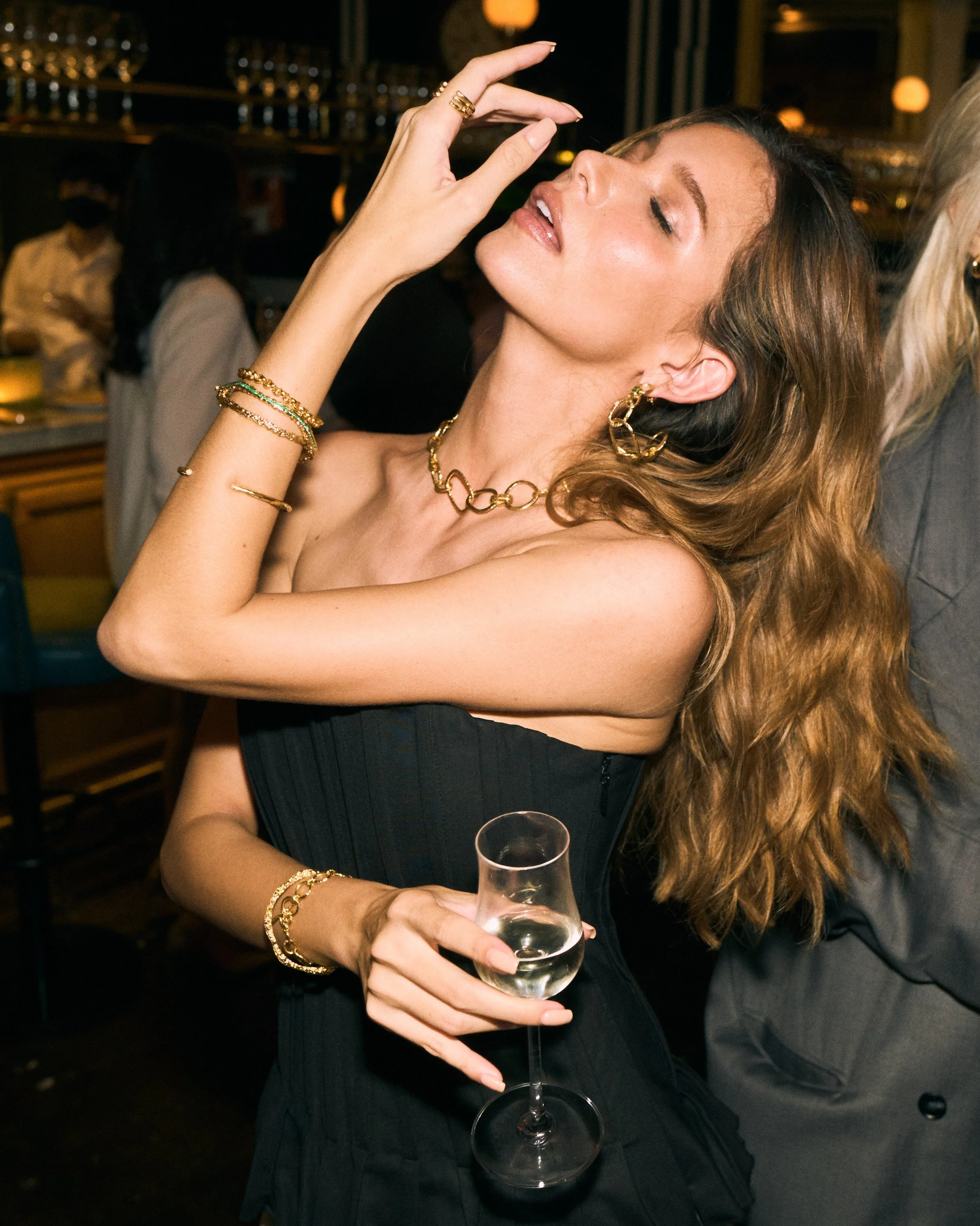 A woman in a black dress holding a glass of white wine at a social gathering in a dimly lit venue, wearing gold jewelry and with long, curly hair.
