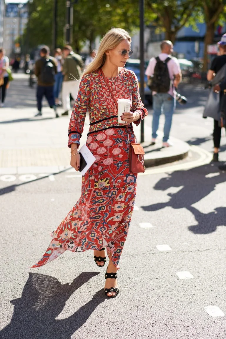 Woman in a red patterned dress walking on the street, carrying a drink and a phone, with other pedestrians in the background.