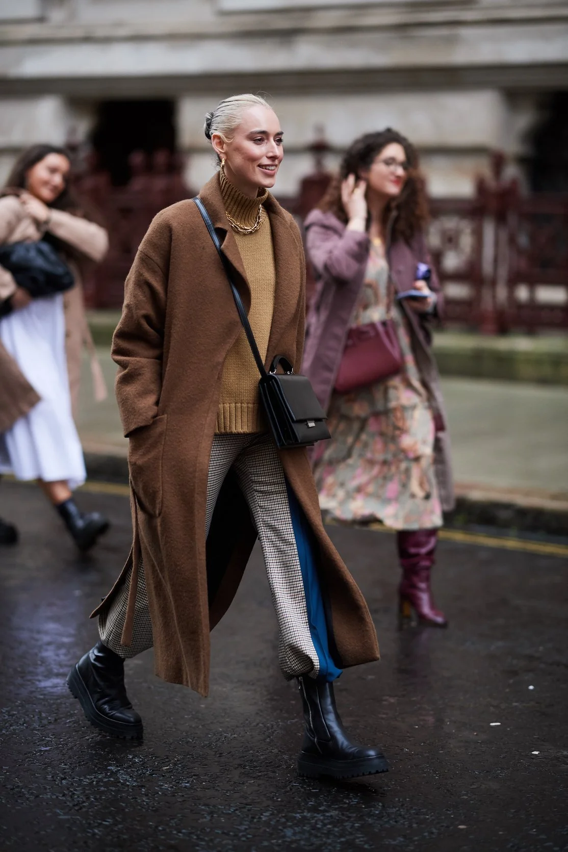 A woman with platinum blonde hair walking outdoors in a city street, wearing a long brown coat, a tan sweater, patterned pants, black boots, and carrying a black crossbody bag. Other women are walking in the background.