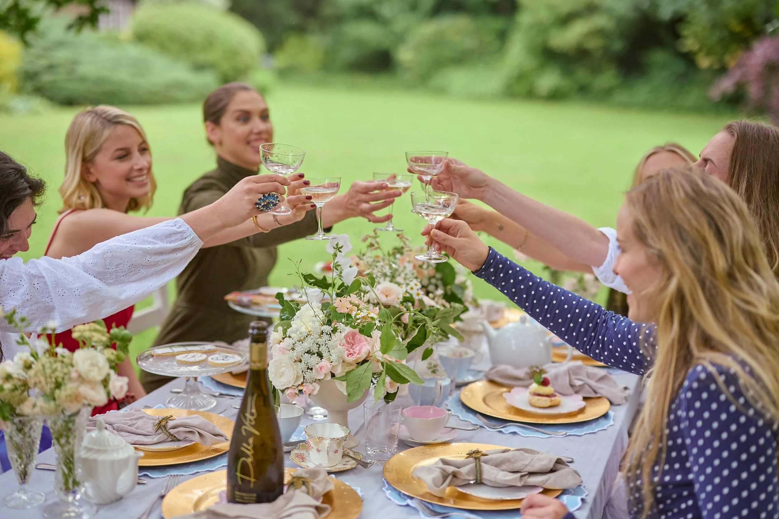 People raising glasses in celebration at a garden party with a decorated table, flowers, and outdoor greenery.