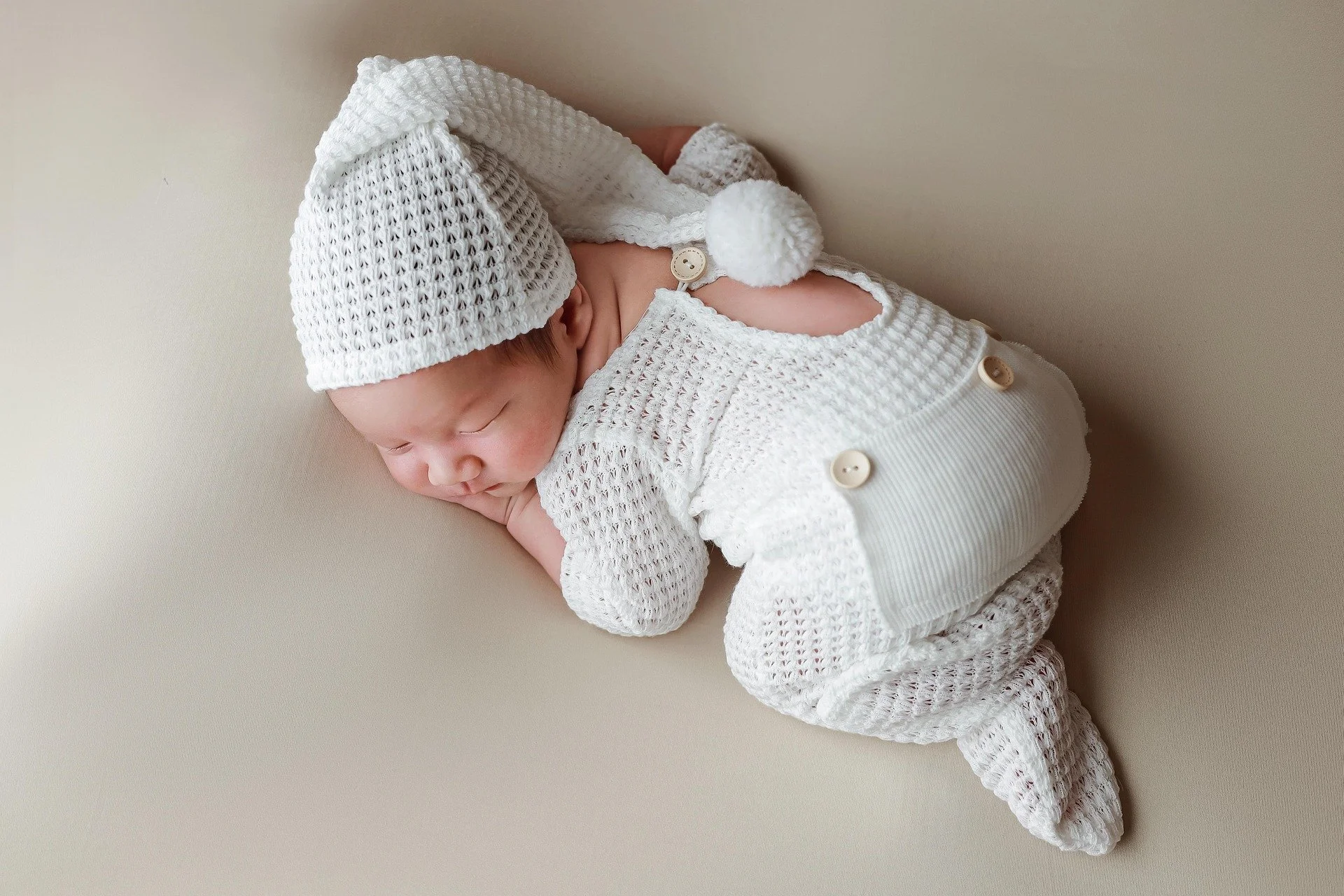 Sleeping newborn wearing white knit pom-pom nightcap and cozy overalls Gentle Night