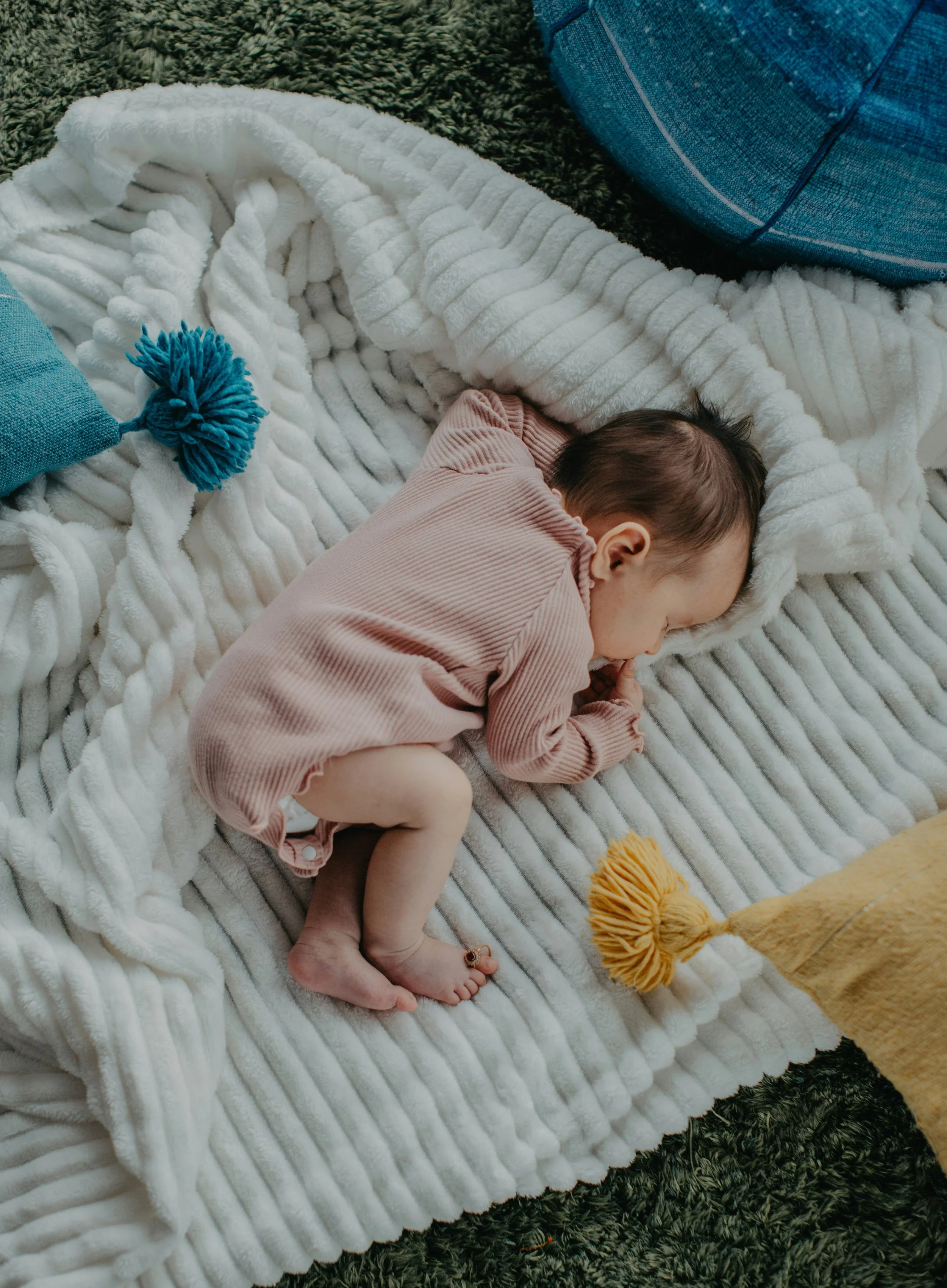 A young child lying peacefully on a plush white blanket on a dark shaggy rug, with children's toys nearby.