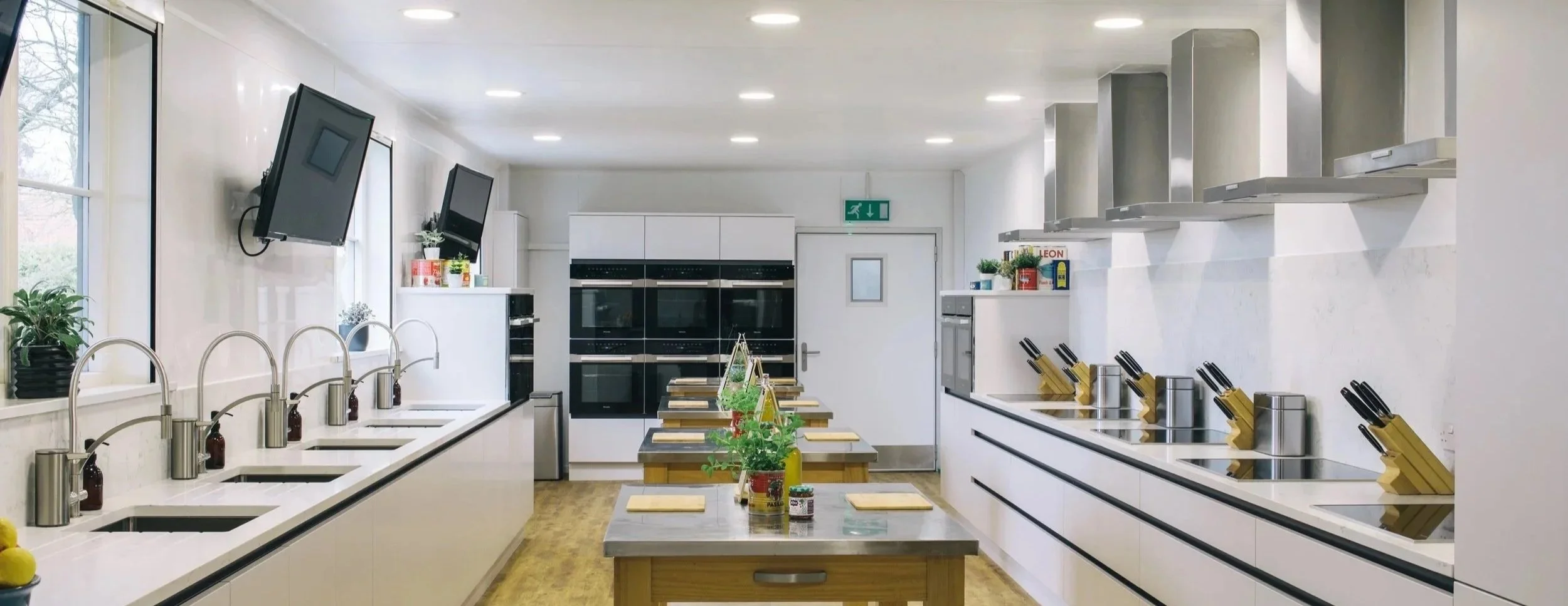 A modern kitchen with white walls, a long countertop with sinks on the left, and multiple stovetops with knife sets on the right. There are two TVs mounted on the wall, and plants on the windowsills. Several wooden tables with potted plants and jars are in the center of the room.