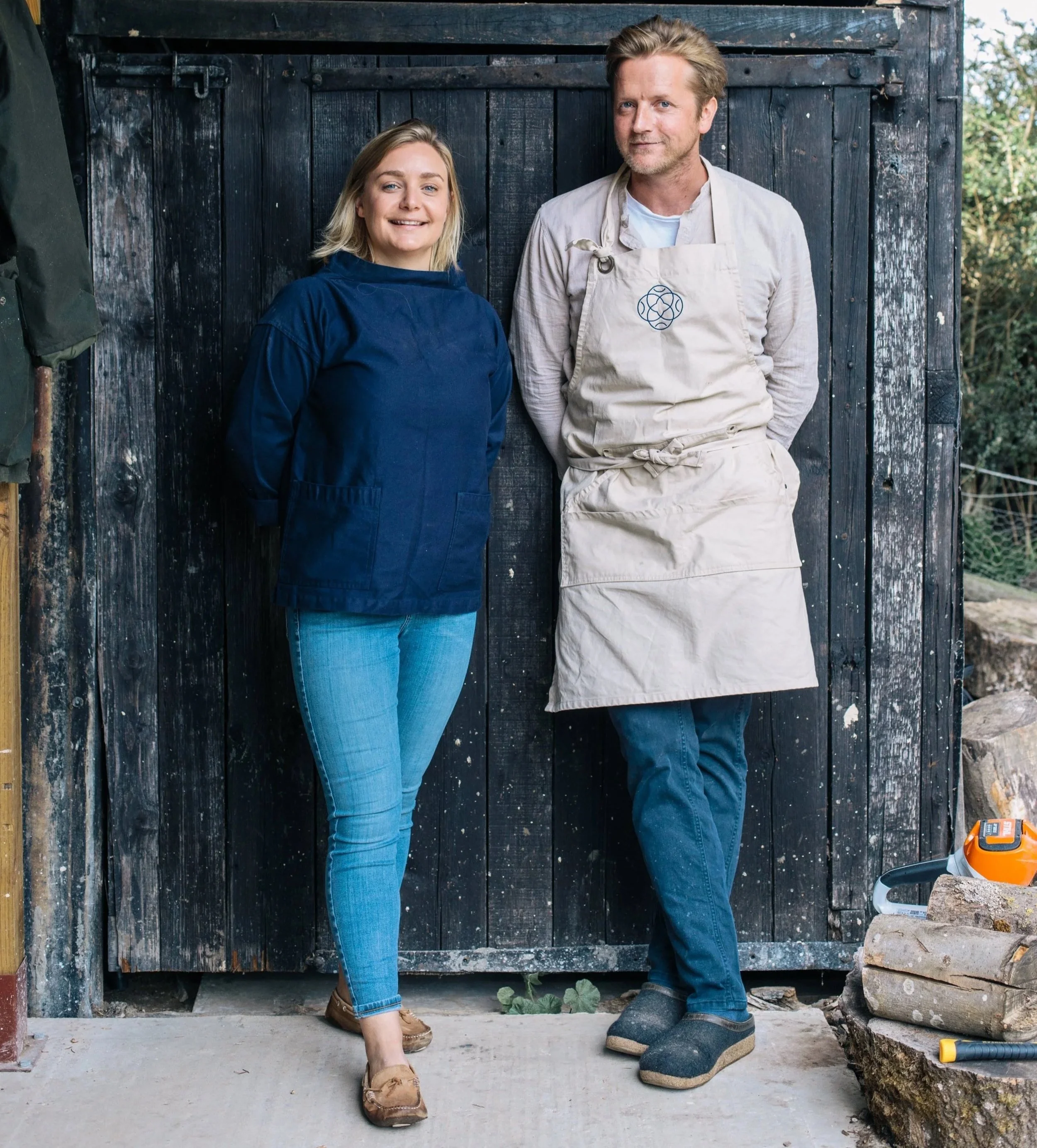 A woman and a man standing side by side outdoors, leaning against a black wooden shed. The woman has blonde hair, is smiling, and wearing a dark blue work shirt and jeans. The man has light brown hair, a beard, and is dressed in a beige apron over a long sleeve shirt and jeans. Behind them are trees and outdoor equipment, including log cutting tools and wood logs.