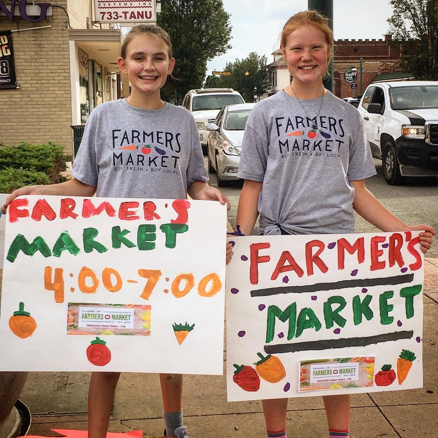 Two girls wearing farmers market t-shirts hold hand-painted signs that say "Farmers Market 4-7pm"
