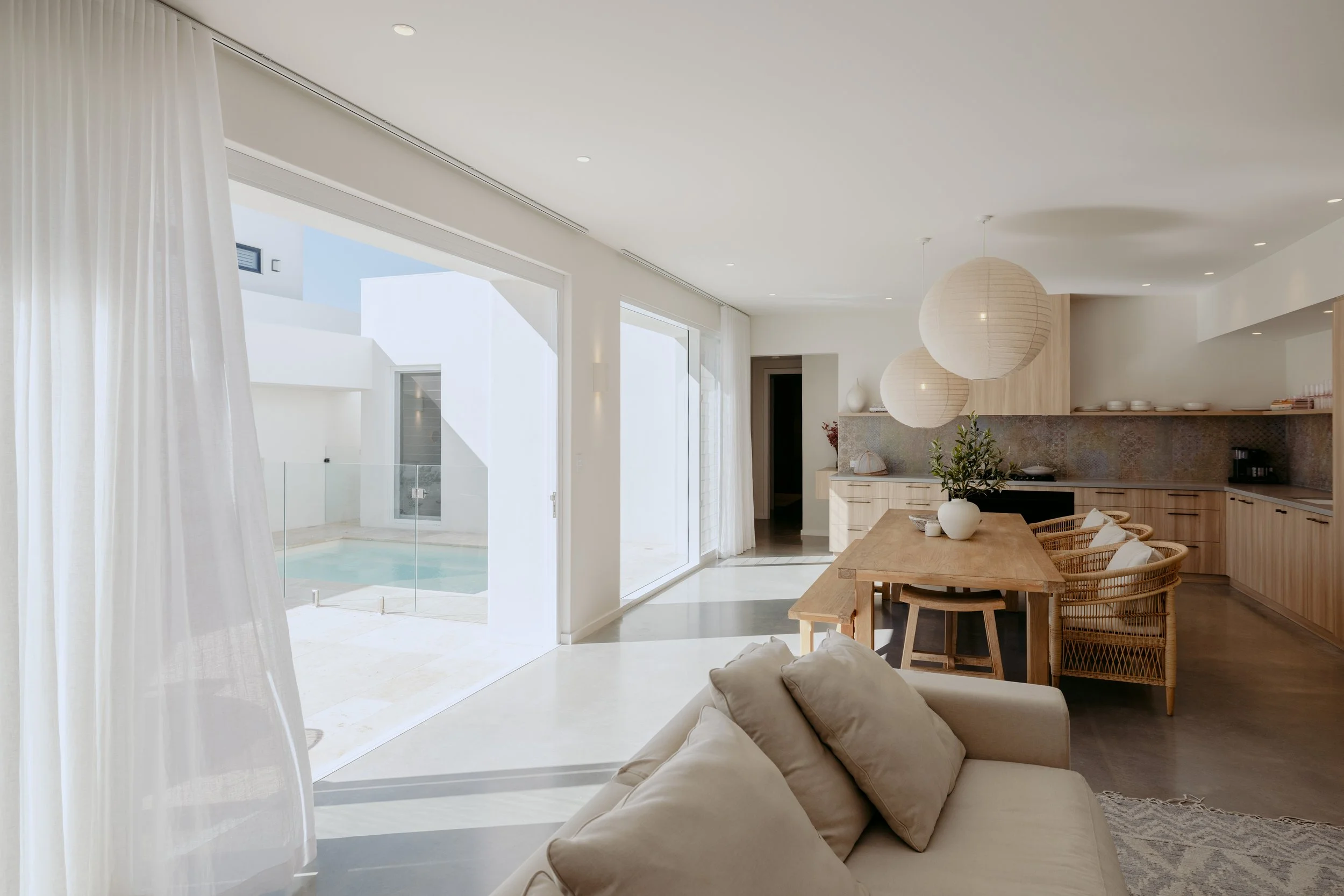 Bright open-concept living and dining area with sliding glass doors leading to a pool area outdoors, beige sectional sofa, wooden dining table with wicker chairs, modern kitchen with light wood cabinets, large paper lanterns hanging from the ceiling,