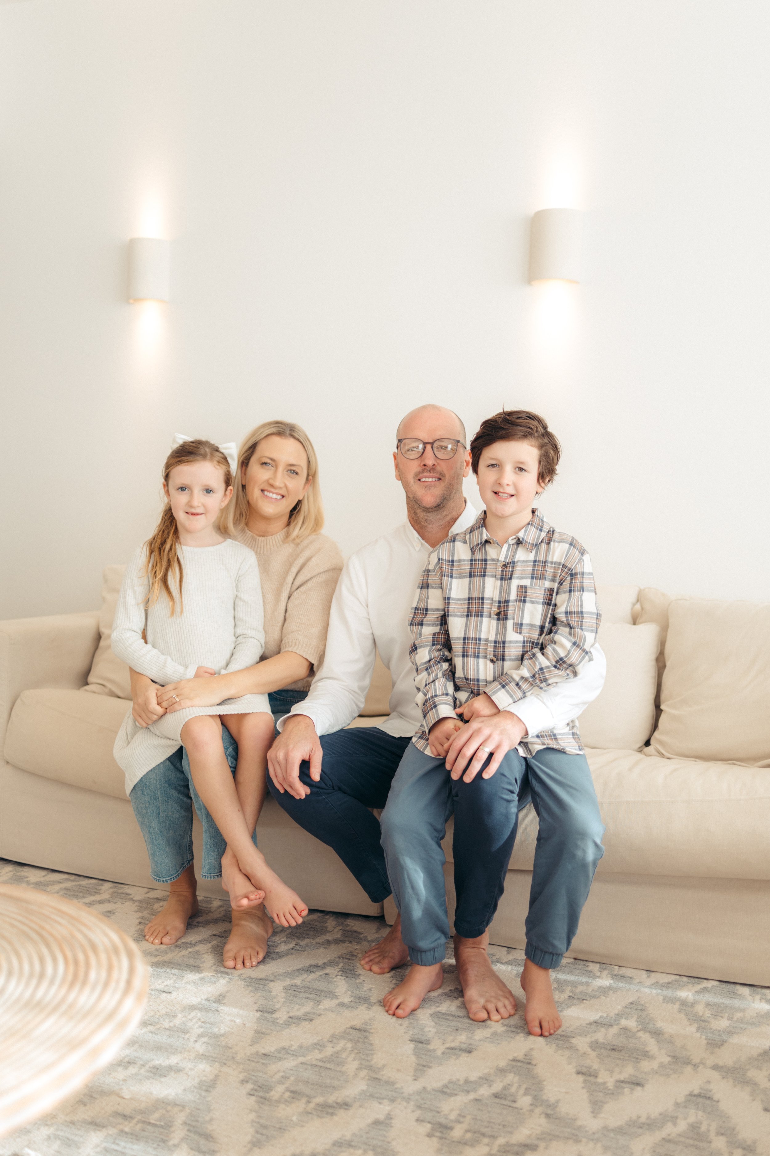 A family of four, including a man, woman, girl, and boy, sitting on a light-colored sofa in a bright, minimalistic living room.