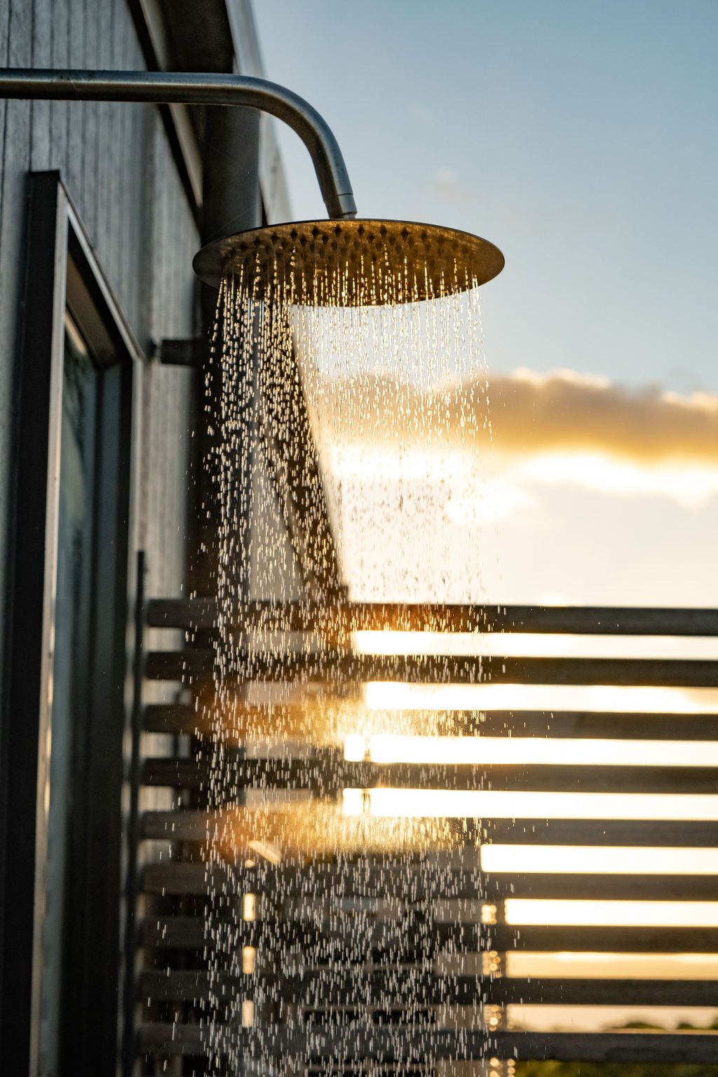 An outdoor shower head releasing water during sunset with a wooden wall and a sunset sky in the background.