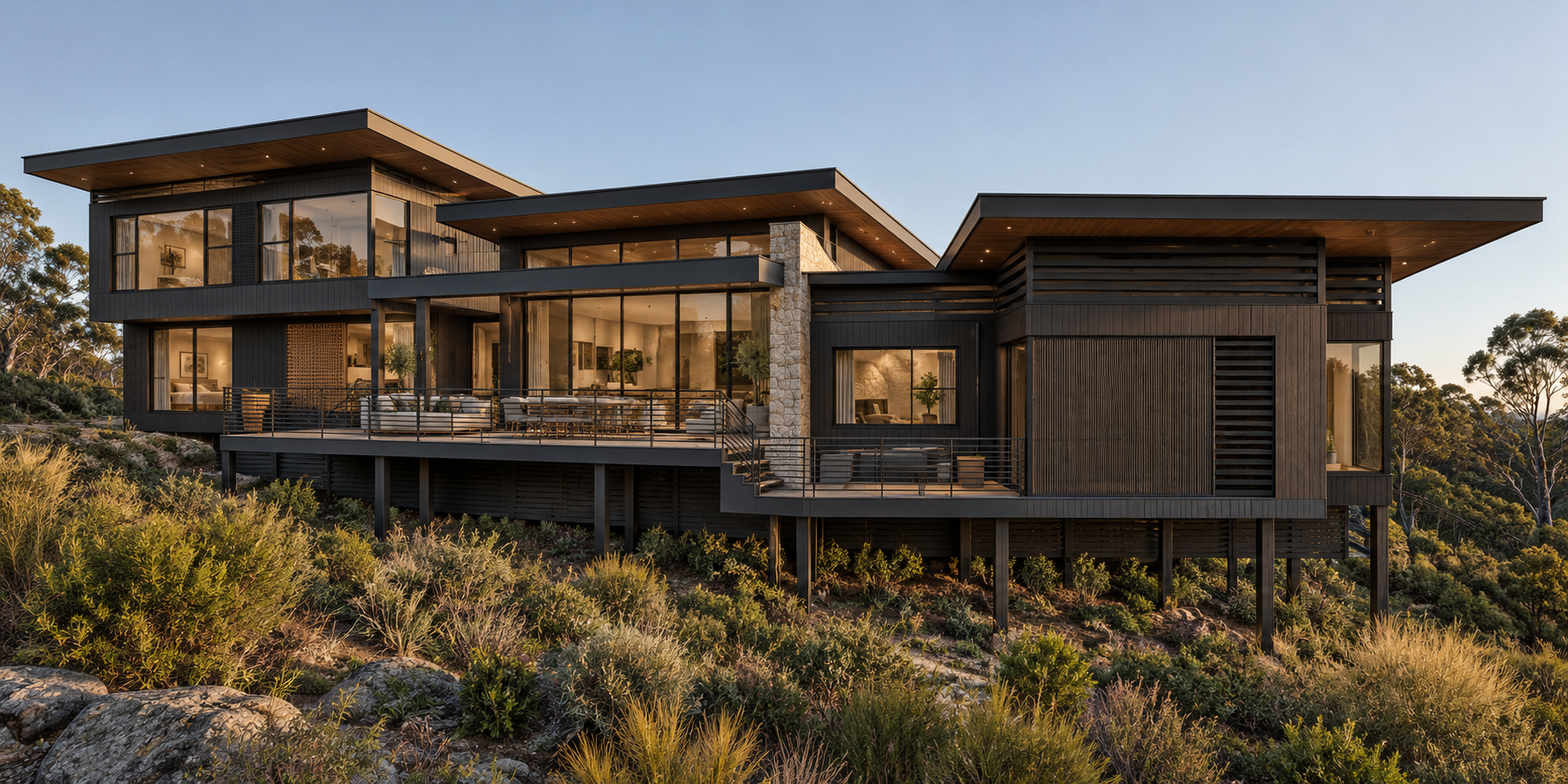 Modern multi-story house on a hill with large glass windows, surrounded by natural vegetation, under a clear sky.