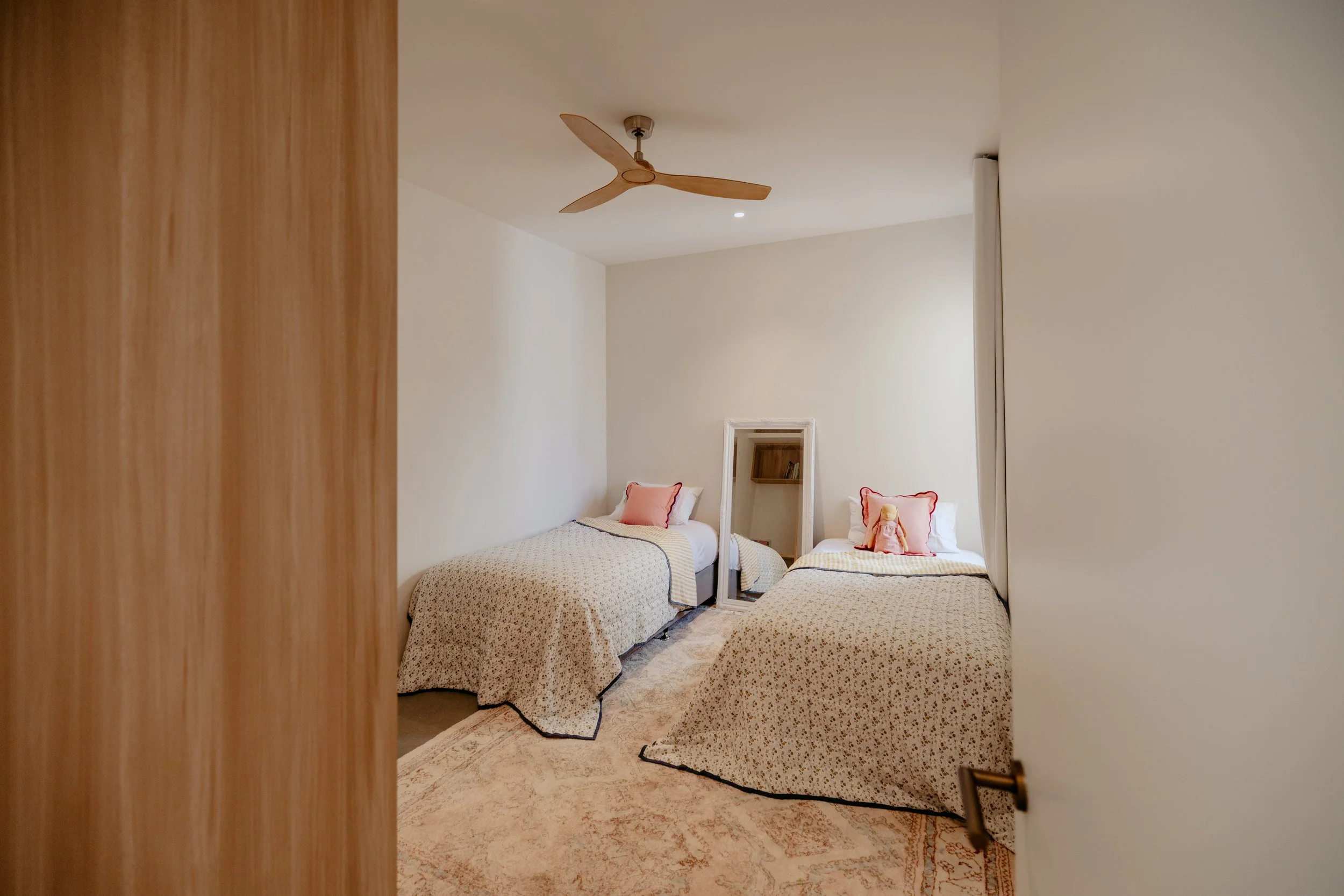 A bedroom with two single beds, one with a pink doll and both with pink pillows, wooden ceiling fan, mirror, and patterned carpet.