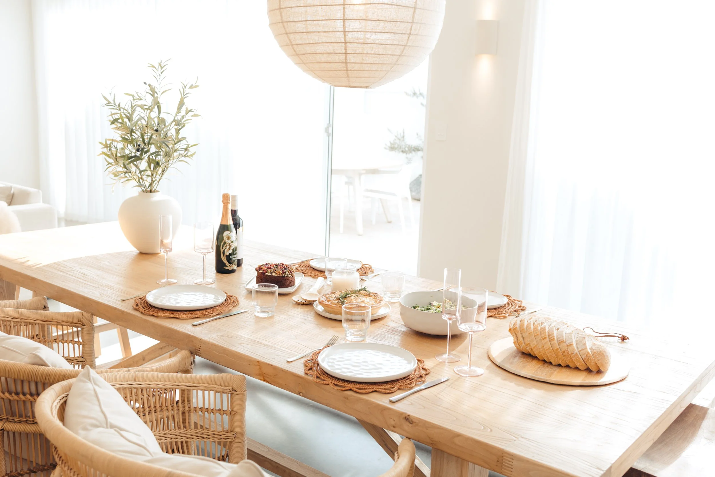 A bright dining room with a light wooden table set for a meal, featuring plates, glasses, a loaf of bread, a bowl of salad, a cake, and bottles of wine, with a white vase of greenery and large paper lantern overhead.