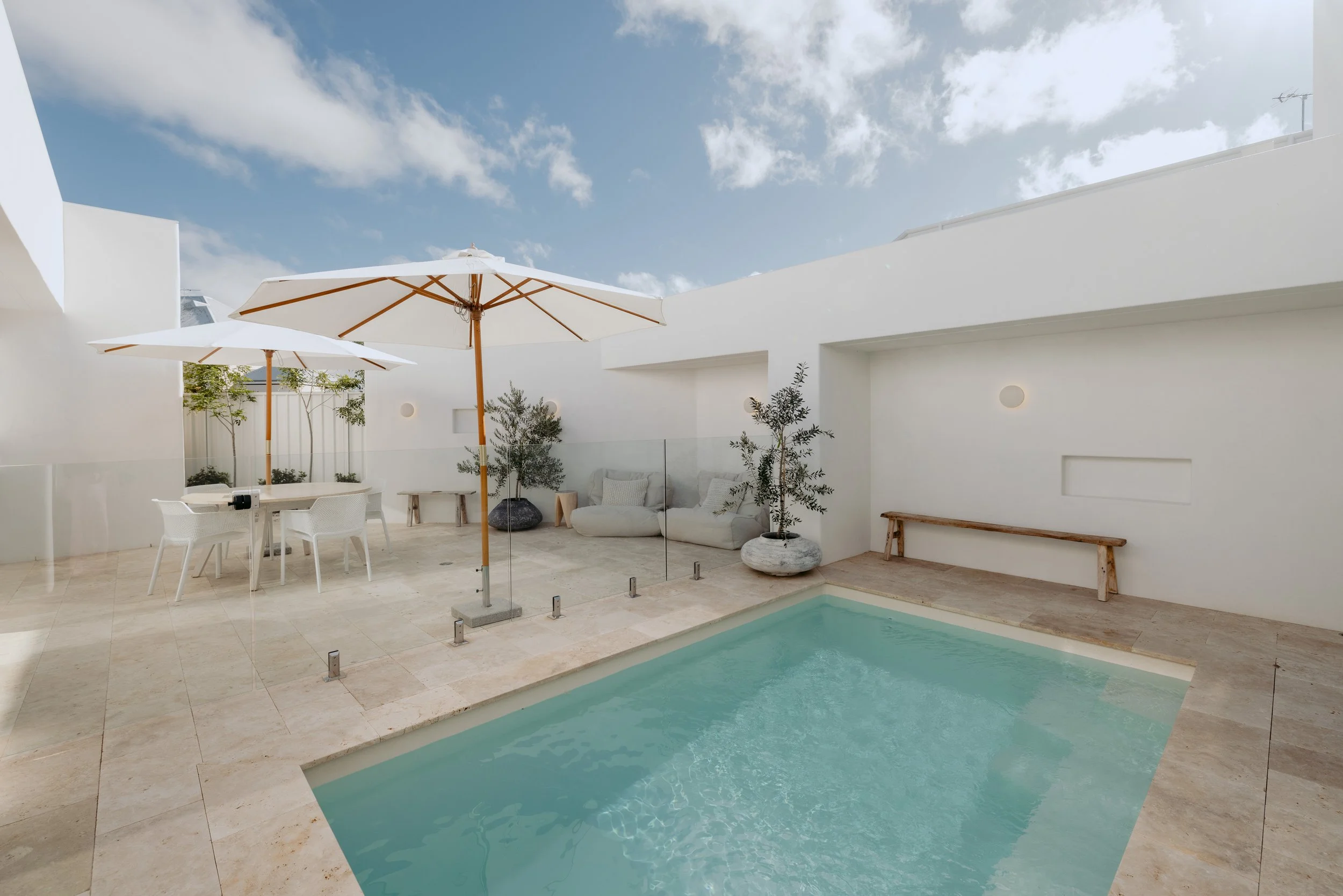 Modern rooftop pool area with white walls, two large white umbrellas, a small table with white chairs, potted plants, a glass fence, a lounge sofa with cushions, and a wooden bench, under a partly cloudy sky.
