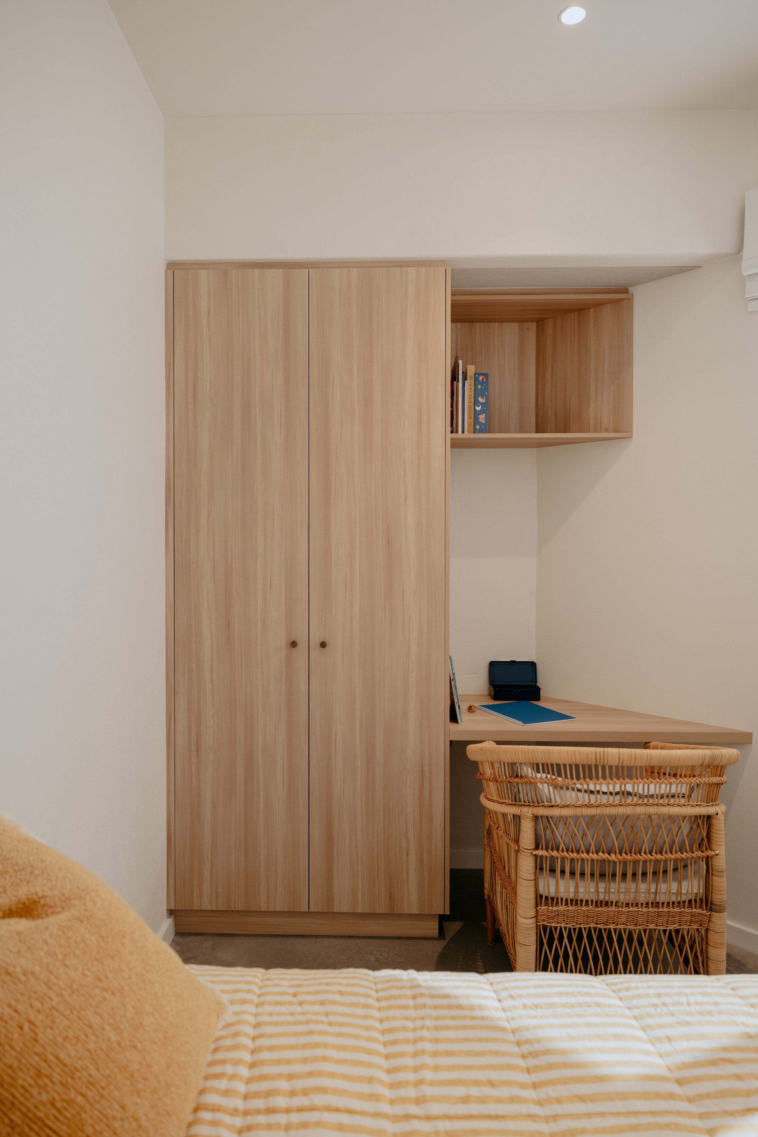 A cozy bedroom corner with a wooden wardrobe, a desk with a blue notebook and a pen, a wicker chair, and a partially visible bed with a yellow pillow and a striped bedspread.