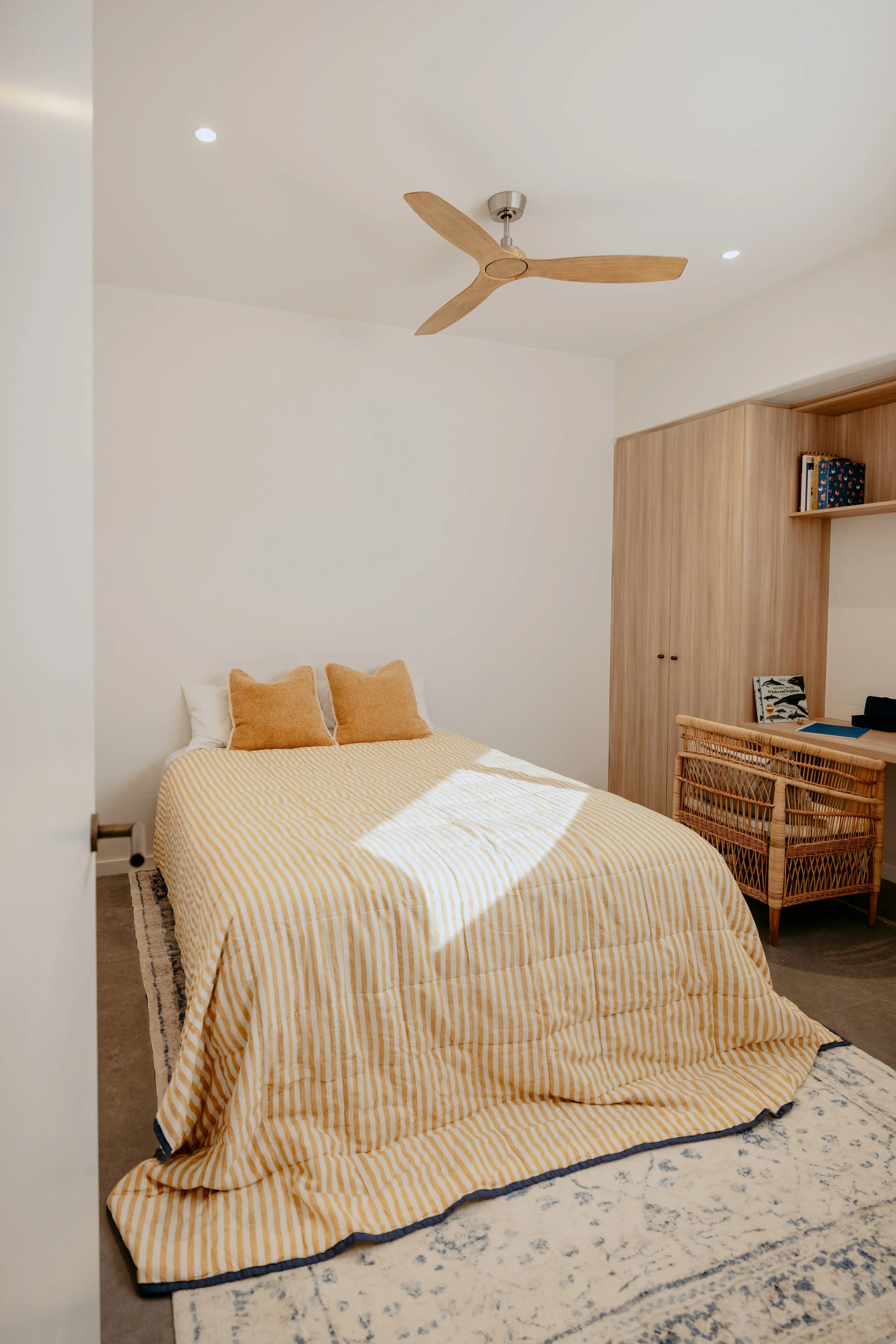 A neatly made bed with yellow striped bedding and two matching pillows in a modern bedroom. The room has a wood wardrobe, a wicker desk, and a ceiling fan.
