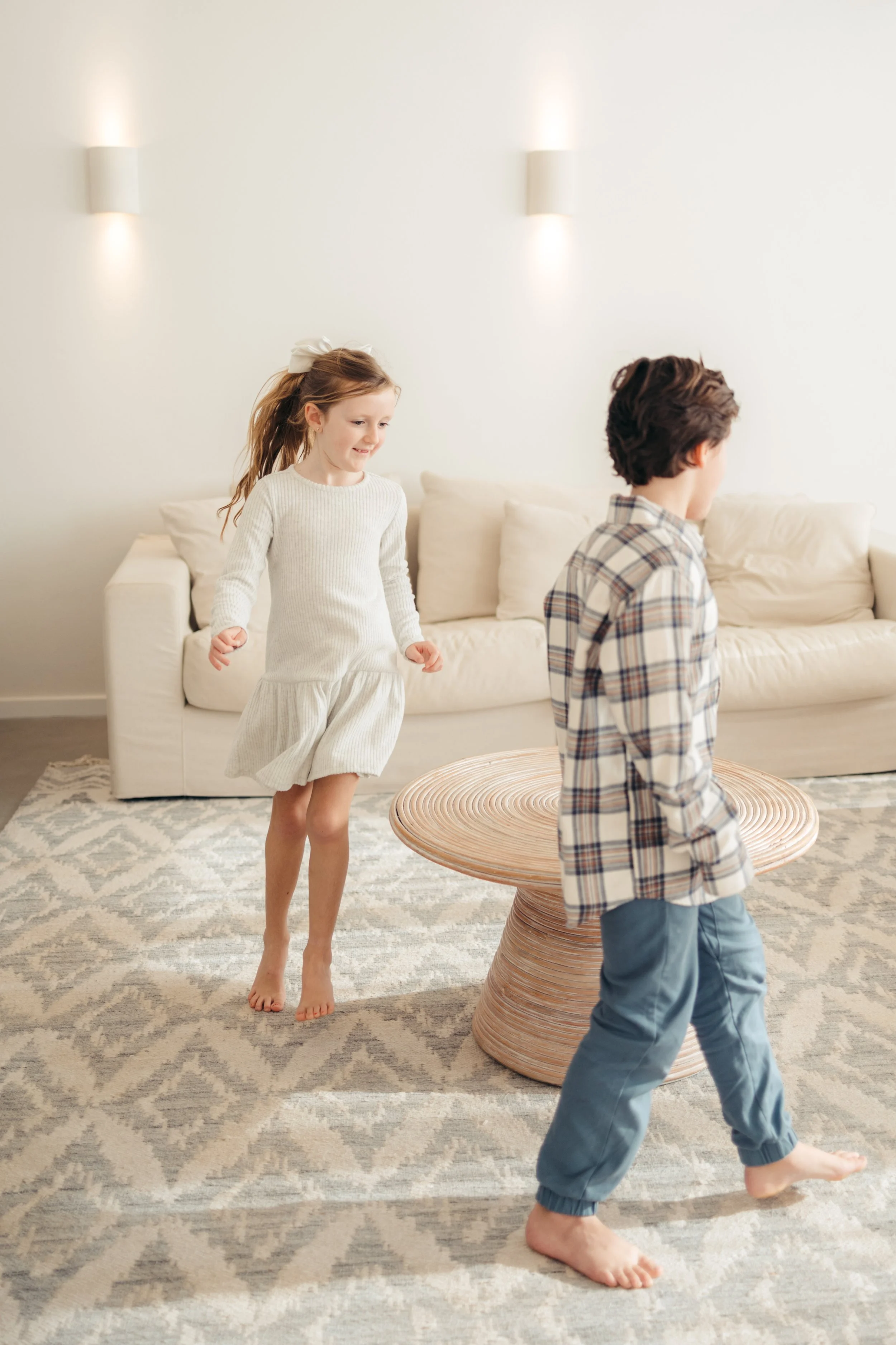 A young girl in a white dress and a boy in a plaid shirt and blue pants playing indoors on a patterned rug, with a beige sofa in the background.