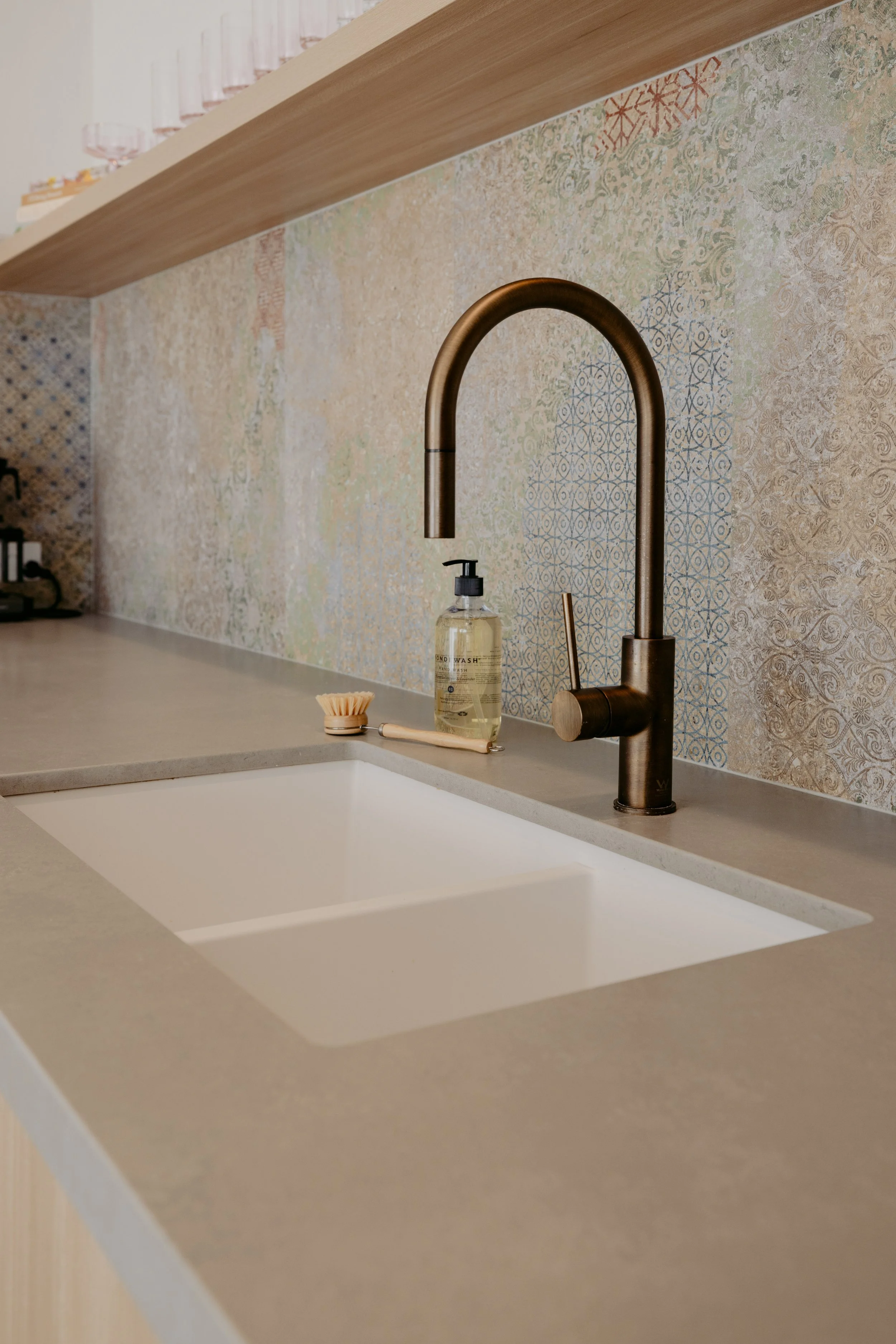 Modern kitchen with a beige countertop, a bronze faucet, a bottle of soap, a scrub brush, and patterned tile backsplash.