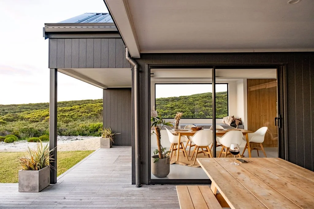 Modern house with deck and large glass sliding doors, offering views of green hills and open landscape, with outdoor potted plants and wooden furniture.