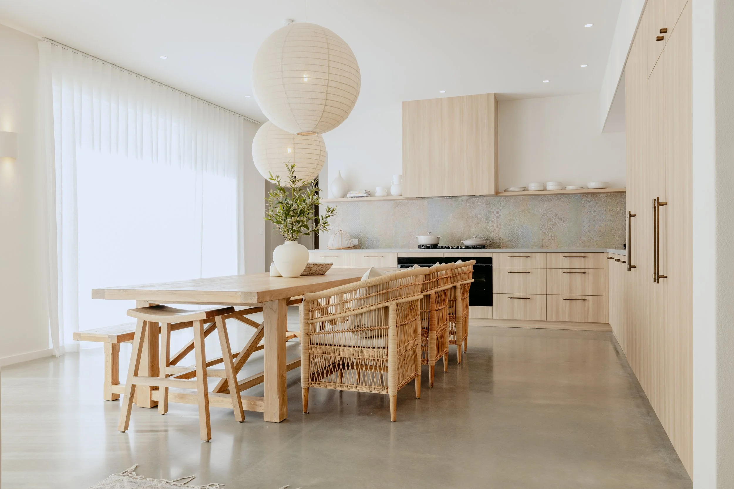 Modern kitchen with light wood cabinets and a dining table with chairs, large paper lanterns, and a potted plant, brightened by natural light from a large sheer-curtained window.