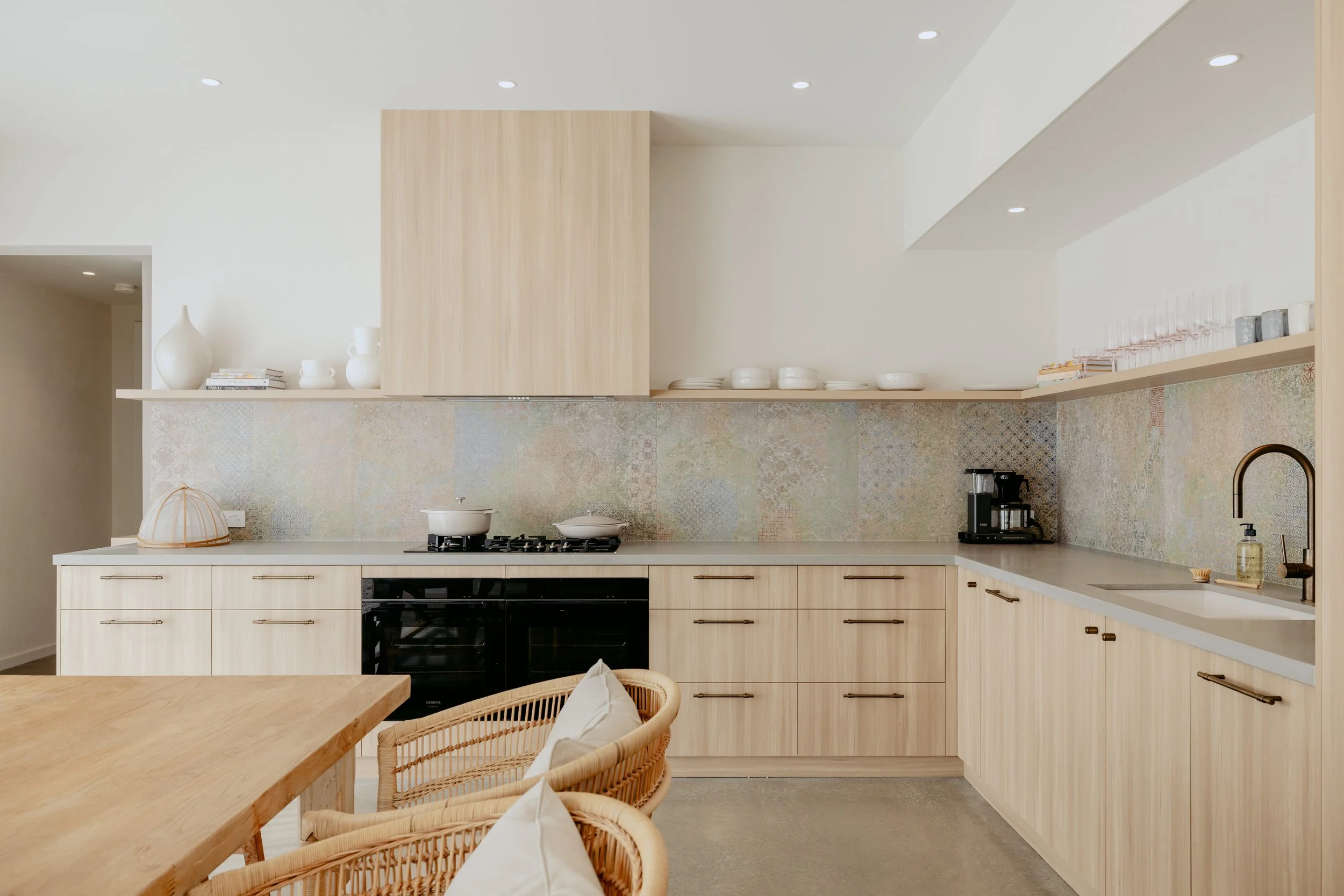 Modern minimalist kitchen with light wood cabinets, beige countertops, and a pastel tile backsplash. There are dishes, glasses, a coffee machine, and a sink with soap. A wooden dining table and wicker chairs are in the foreground.