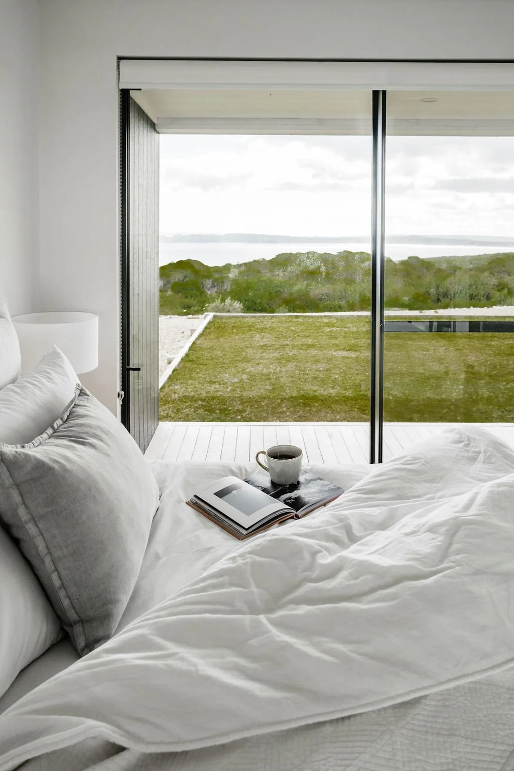 Bedroom with a view of a grassy outdoor area and trees through sliding glass doors, with an open book and a cup on the bed.