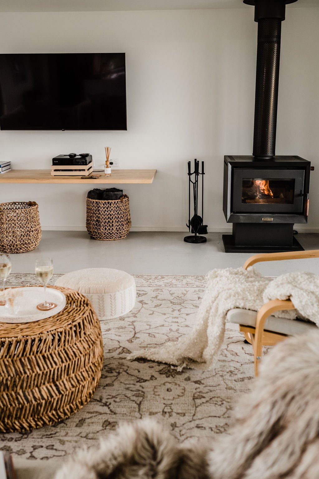 Cozy living room with a wood stove, TV mounted on the wall, woven baskets, a rattan chair with a blanket, a patterned rug, and glass of champagne on a rattan coffee table.