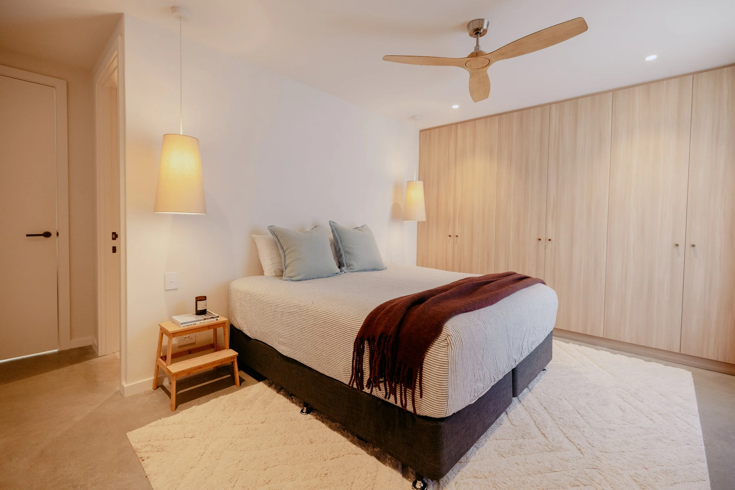 Modern bedroom with a bed, beige bedding, light blue pillows, a dark blanket, wooden side table, beige pendant lamps, wood-clad closet, ceiling fan, white walls, and a cream rug.