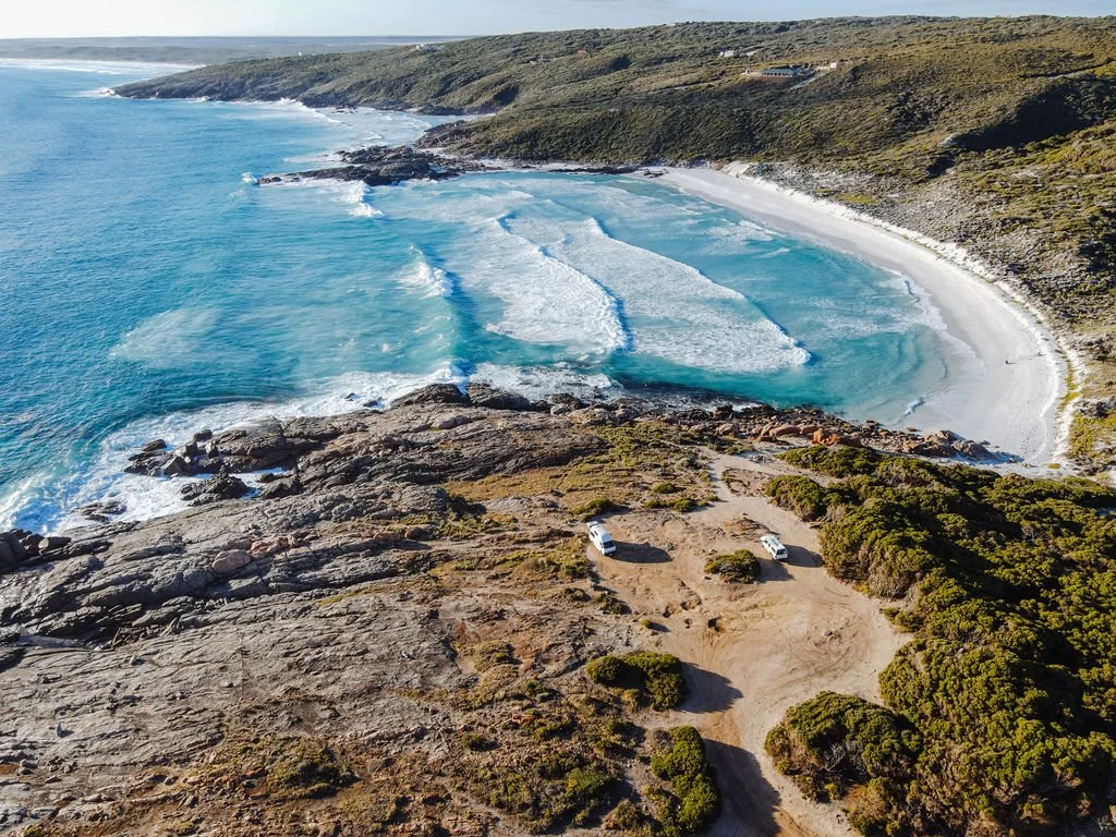 Aerial view of a beach with turquoise ocean waves crashing onto a sandy shoreline, surrounded by rocky cliffs and green shrubbery, with two white vehicles parked on a dirt area.