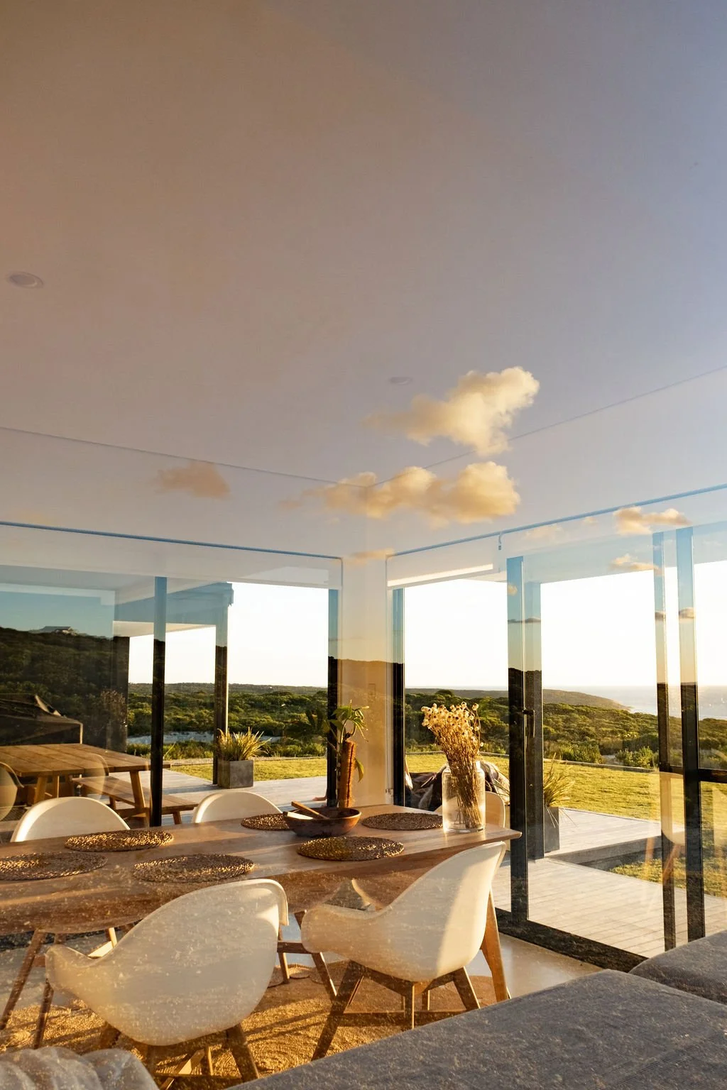 Modern dining room with large glass windows and sliding glass doors, views of green landscape and ocean, decorated with a wooden table, white chairs, woven placemats, and vases with dried flowers, sunlight illuminating the space.
