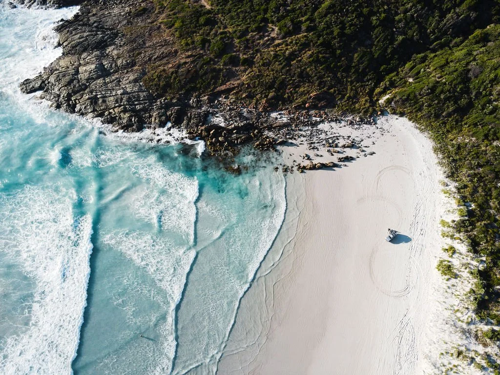Aerial view of a sandy beach with turquoise waves and rocky cliffs covered in green vegetation.