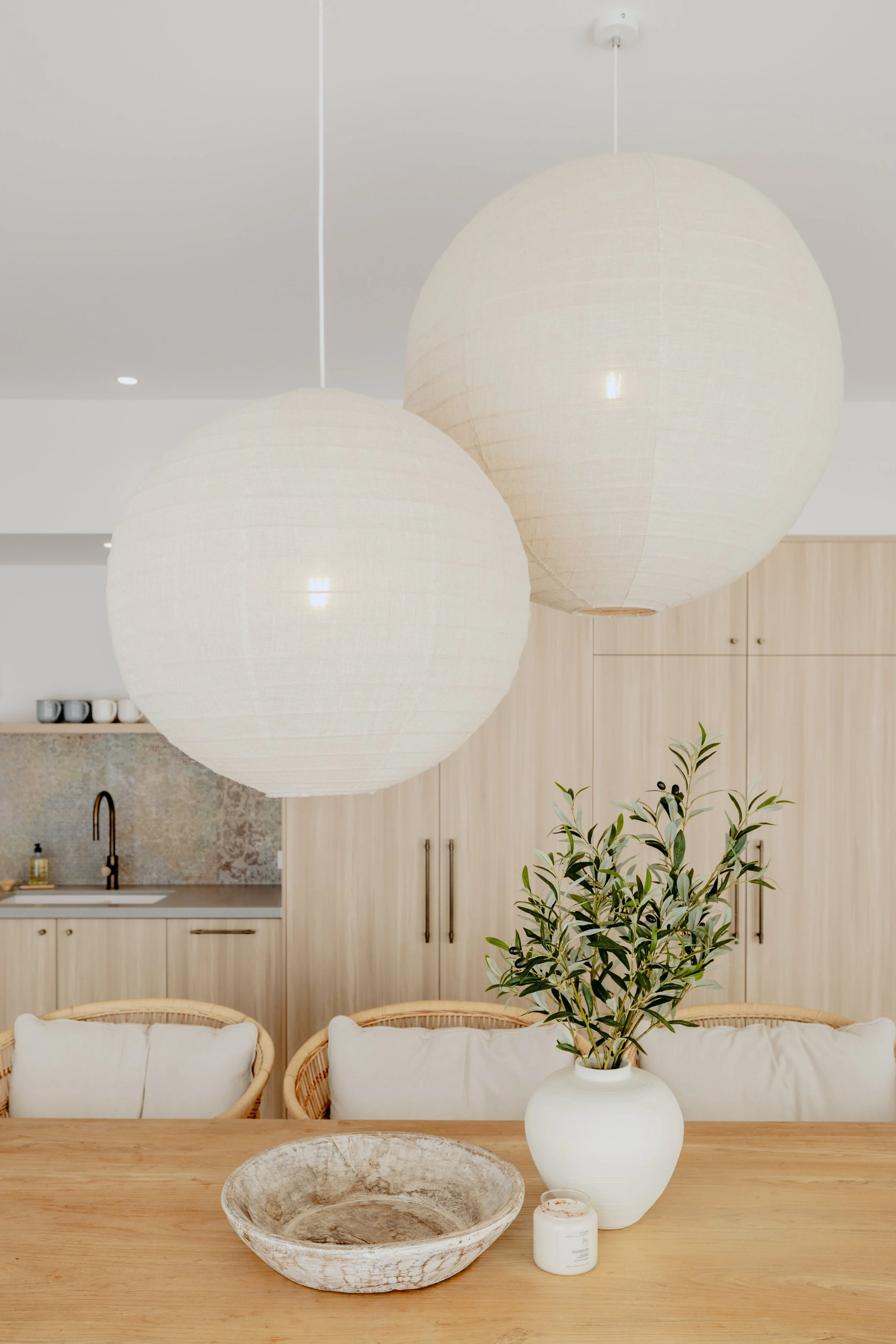Modern kitchen and dining area with two large white paper lantern pendant lights hanging above a wooden dining table. The table has a white vase with green leaves, a round stone bowl, and a small white candle. Cushioned rattan chairs are around the t